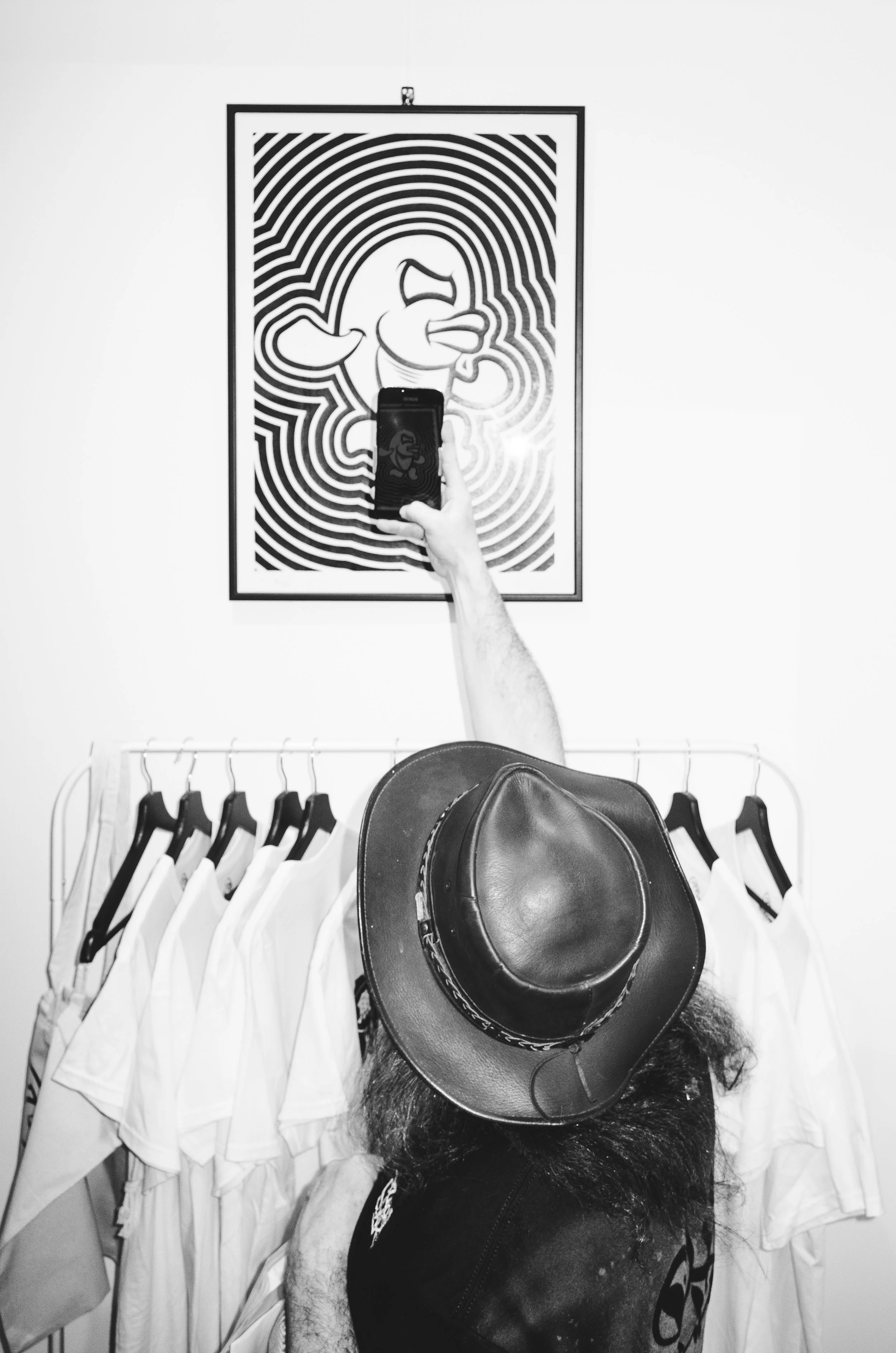 Person taking a selfie in front of a framed art piece, wearing a large hat, with clothes hanging on a rack in the background.