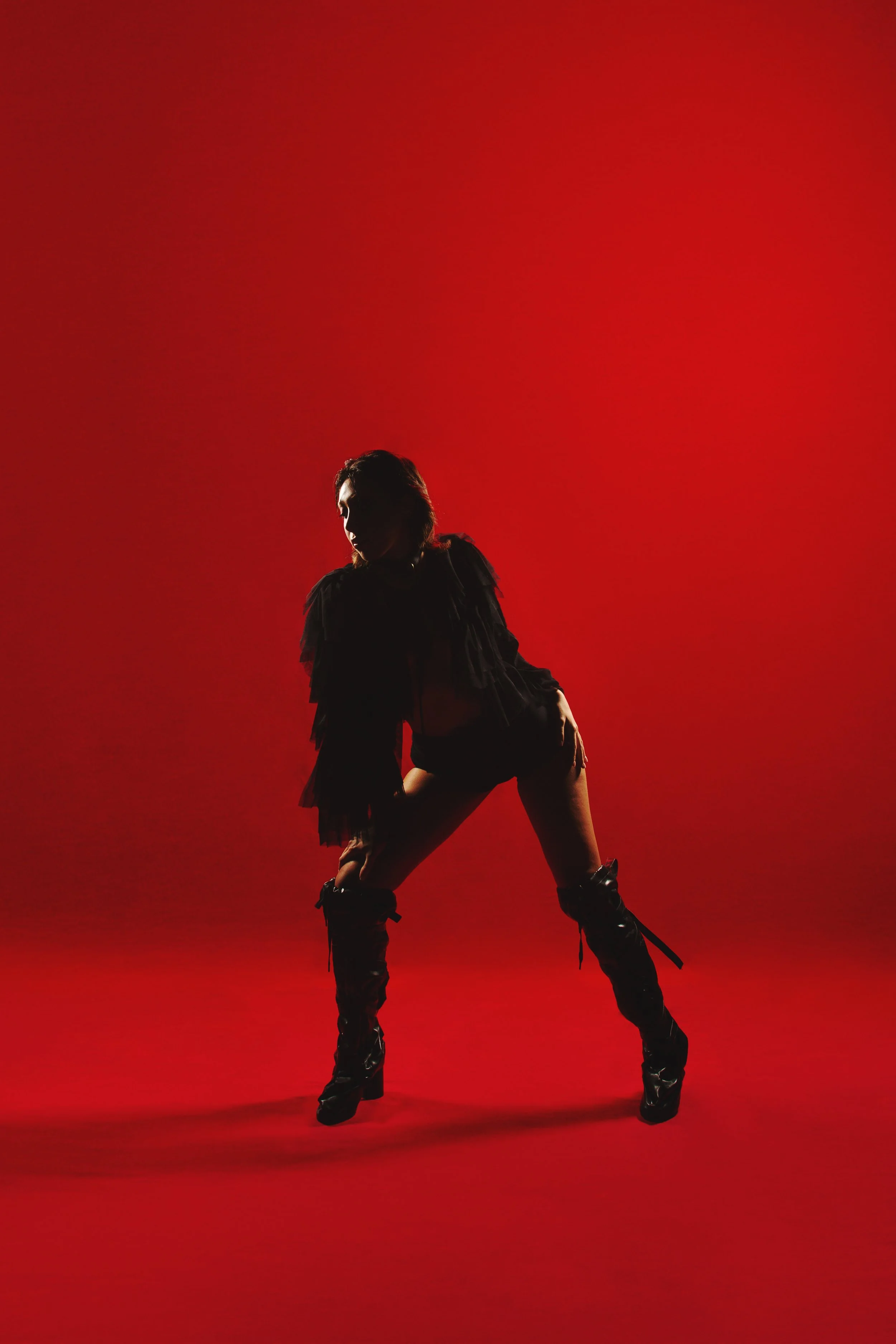 A person in black clothing with tall boots, posing dynamically against a red background with shadowed face.