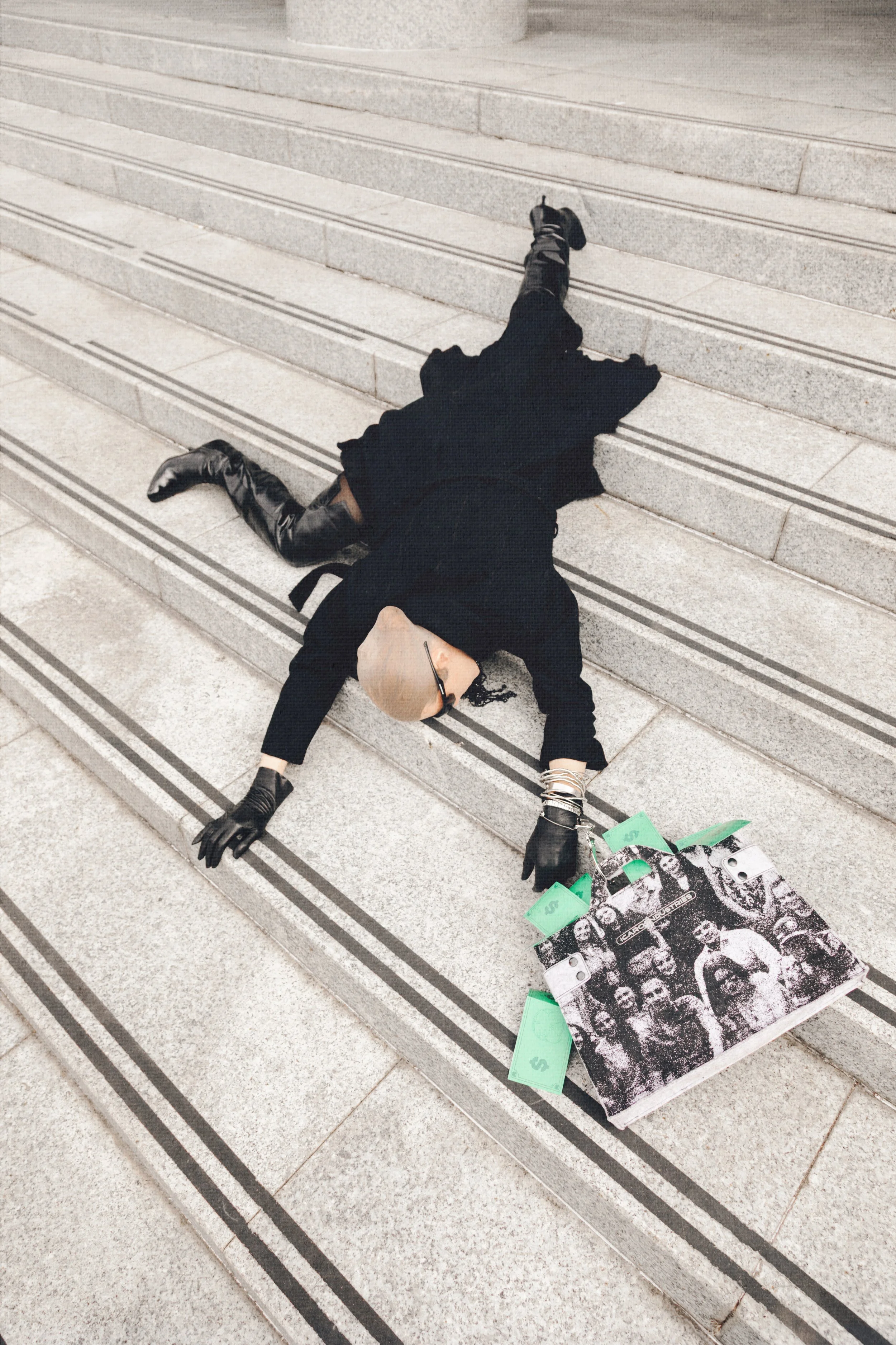 A person dressed in black, with gloves and glasses, lying on a set of stone steps, holding a black-and-white photo collage with green dollar bills attached, in an urban setting.