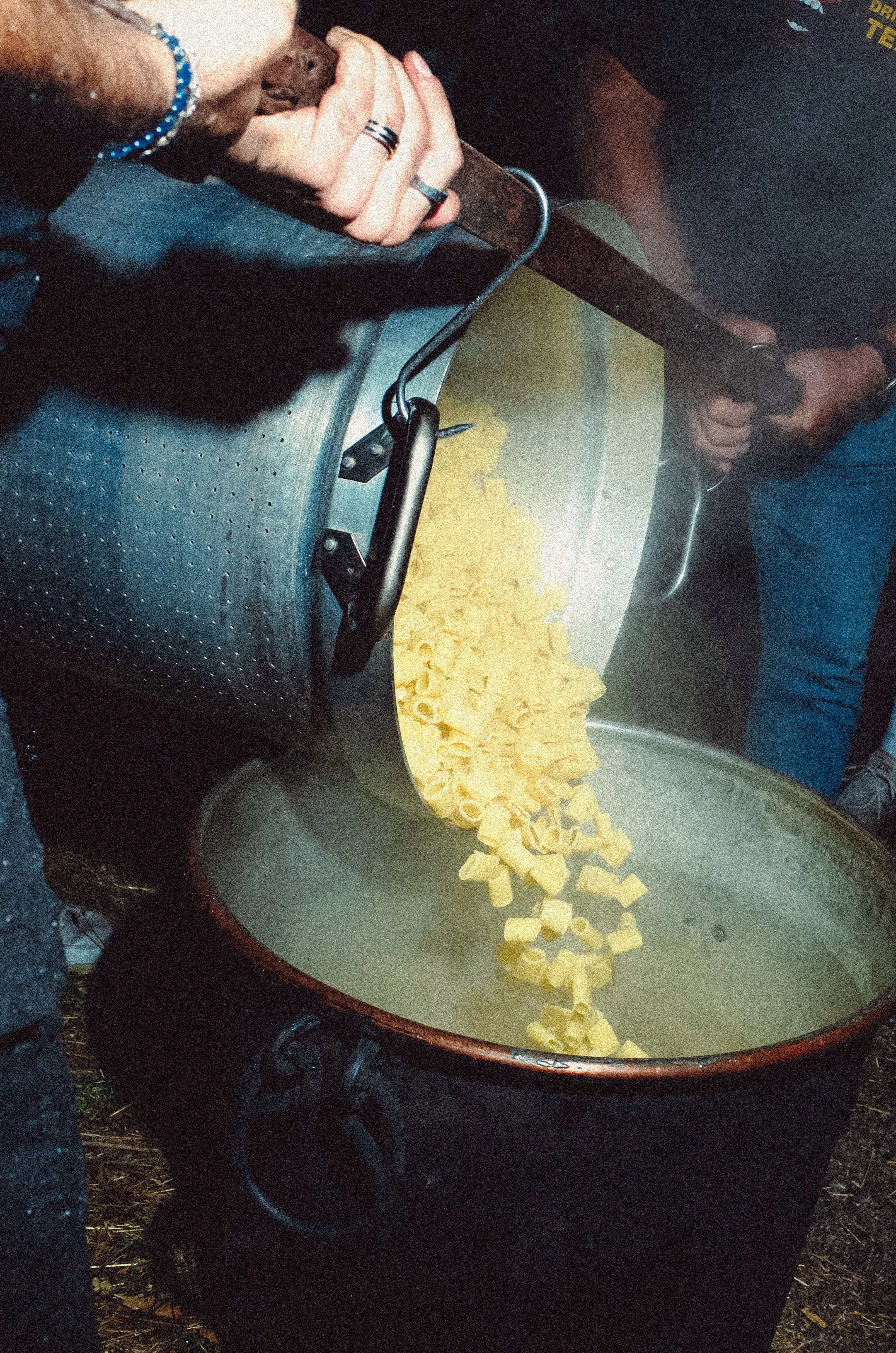 Person pouring cooked pasta from a pot into a large serving bowl outdoors.