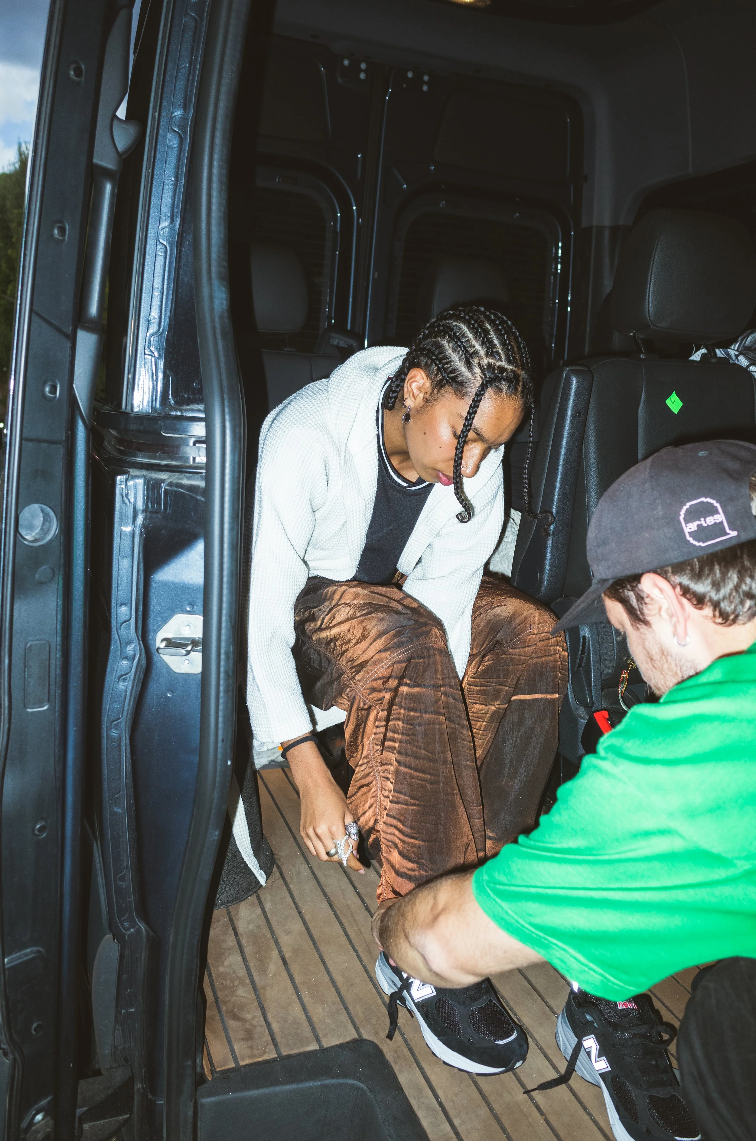 A woman with braided hair sitting in the back of a vehicle, getting her shoe tied by a man wearing a green shirt and a black cap.