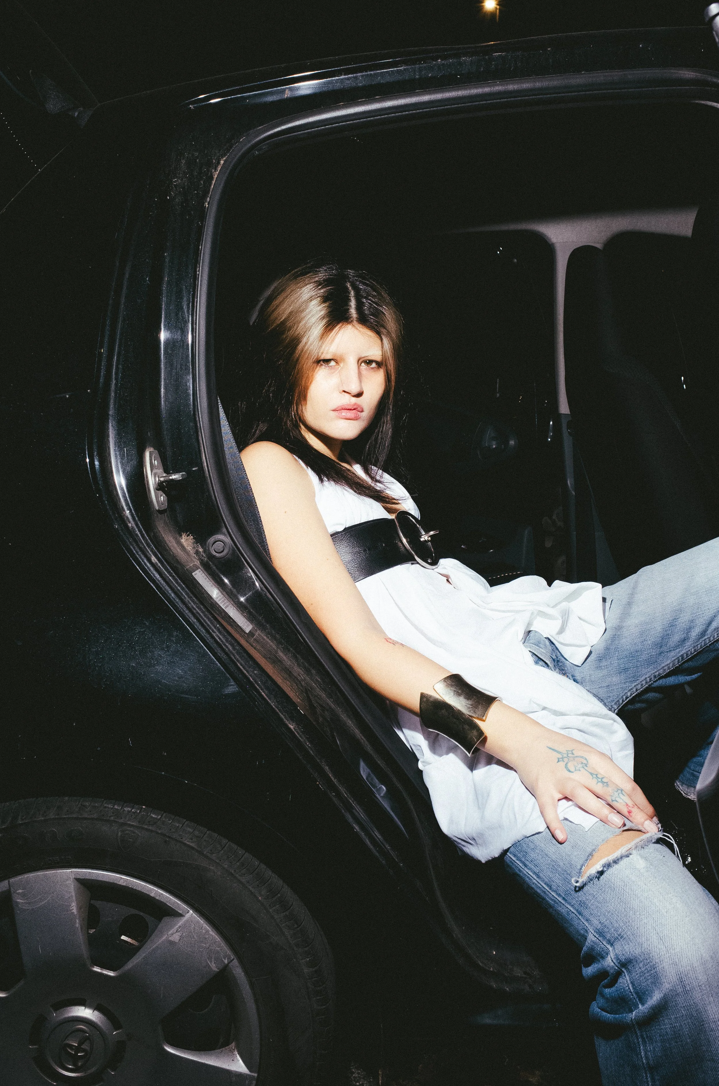 A young woman with dark hair and light highlights sitting in the backseat of a black car at night. She is wearing a white dress, ripped jeans, and gold arm cuffs, looking at the camera with a serious expression.
