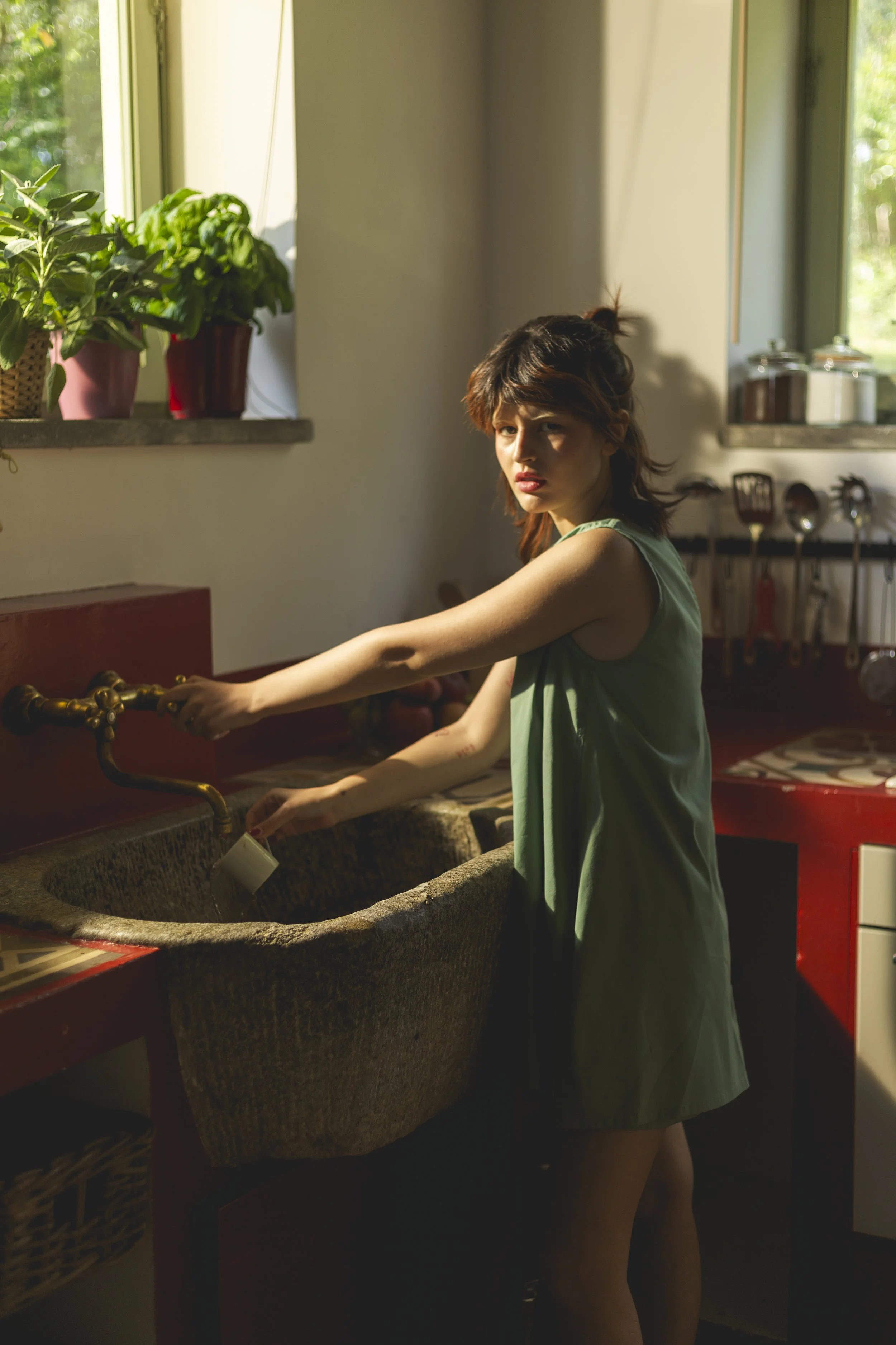A young woman with short, tousled hair and a green sleeveless dress stands at a rustic kitchen sink, holding a white mug under a brass faucet. The sunlight streams through a window with potted plants, illuminating the scene and casting shadows.