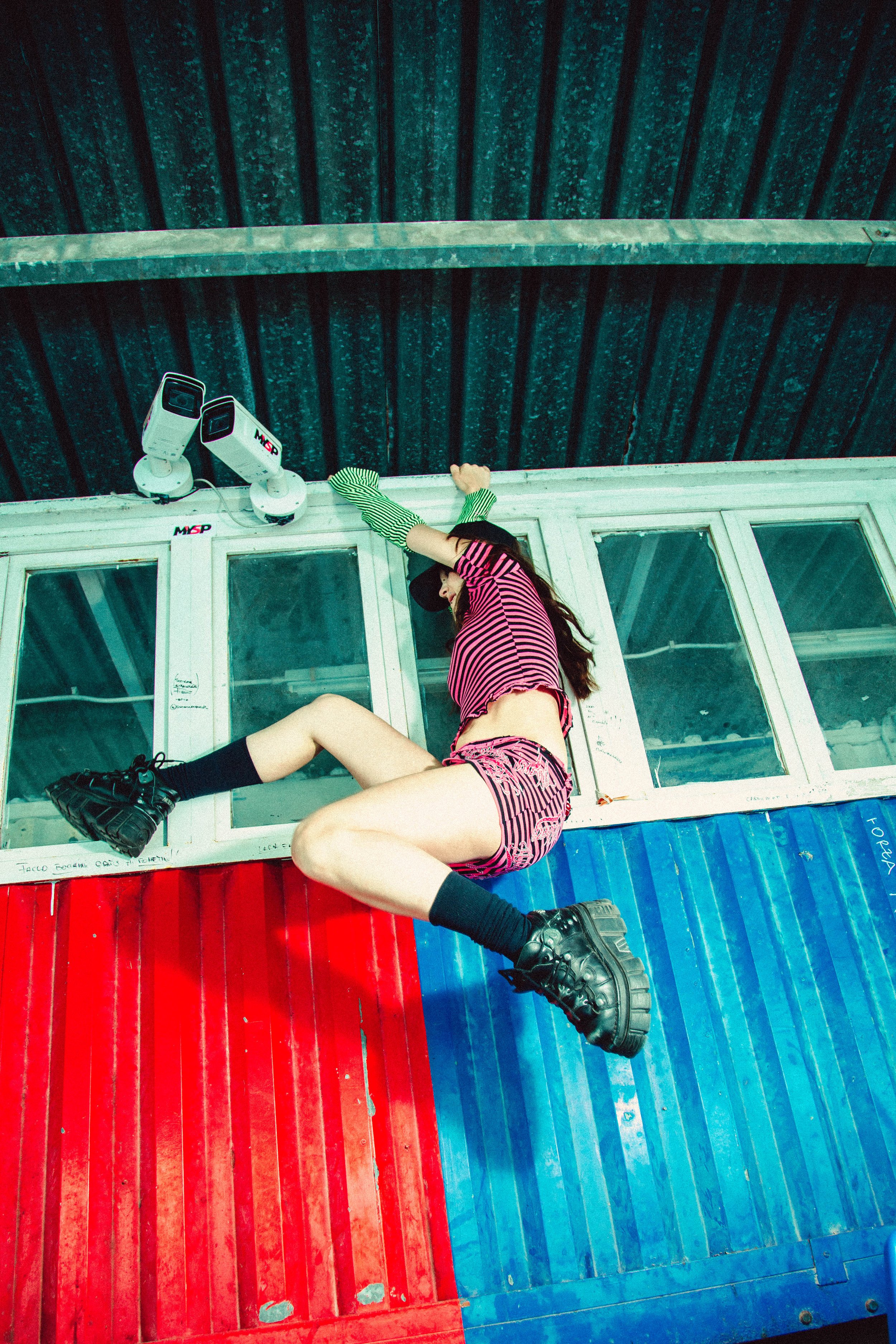 A person with long hair, wearing a pink and black striped shirt, black hat, shorts, and chunky black shoes, lying upside down on a multicolored wall with blue and red panels, next to a window with security cameras above.