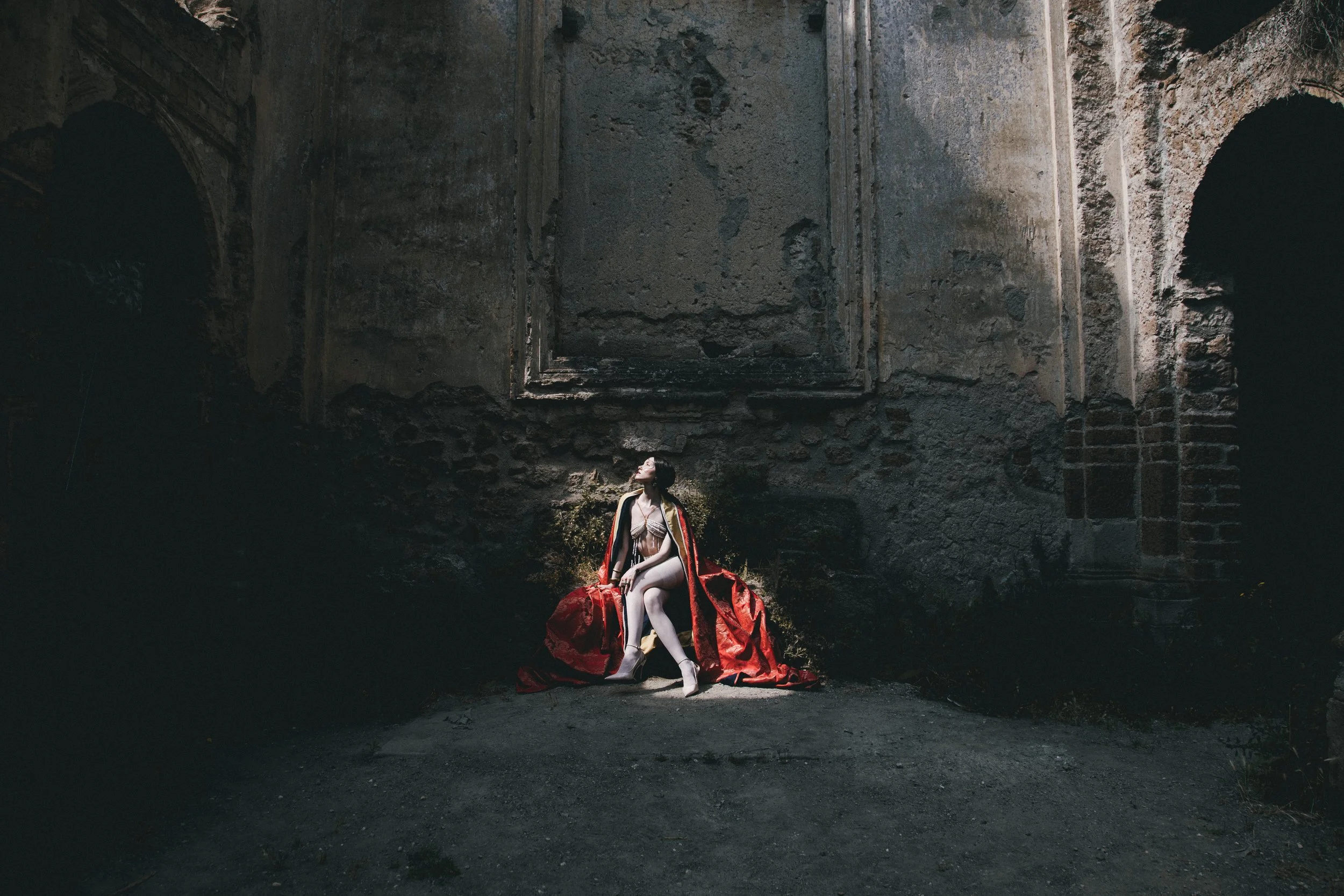 A woman in lingerie and white stockings sitting on a red cape, in a dark, abandoned building with peeling walls and arches.