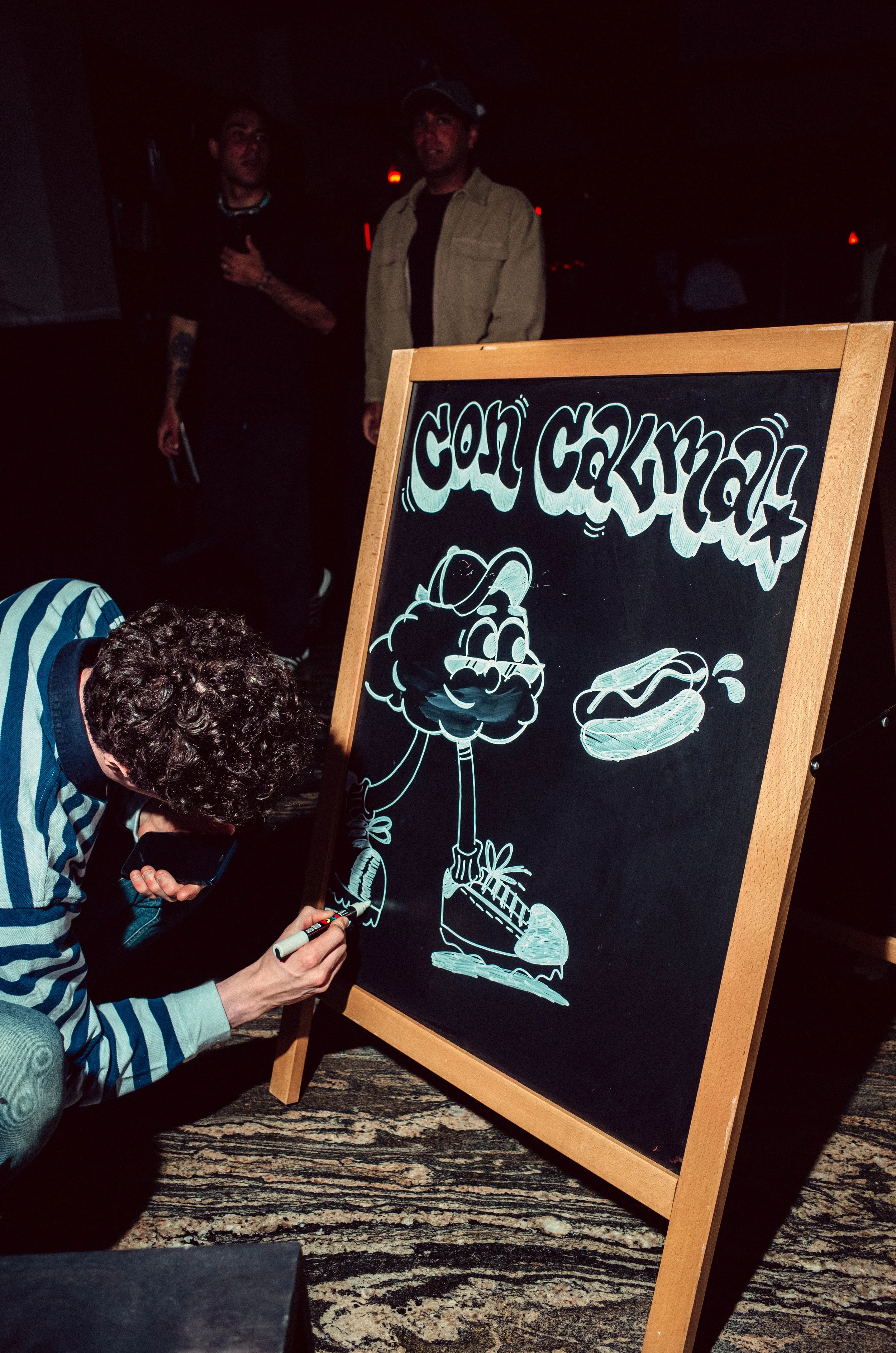 A person drawing on a blackboard sign in a dark venue with a cartoon character and text, with two people standing in the background.