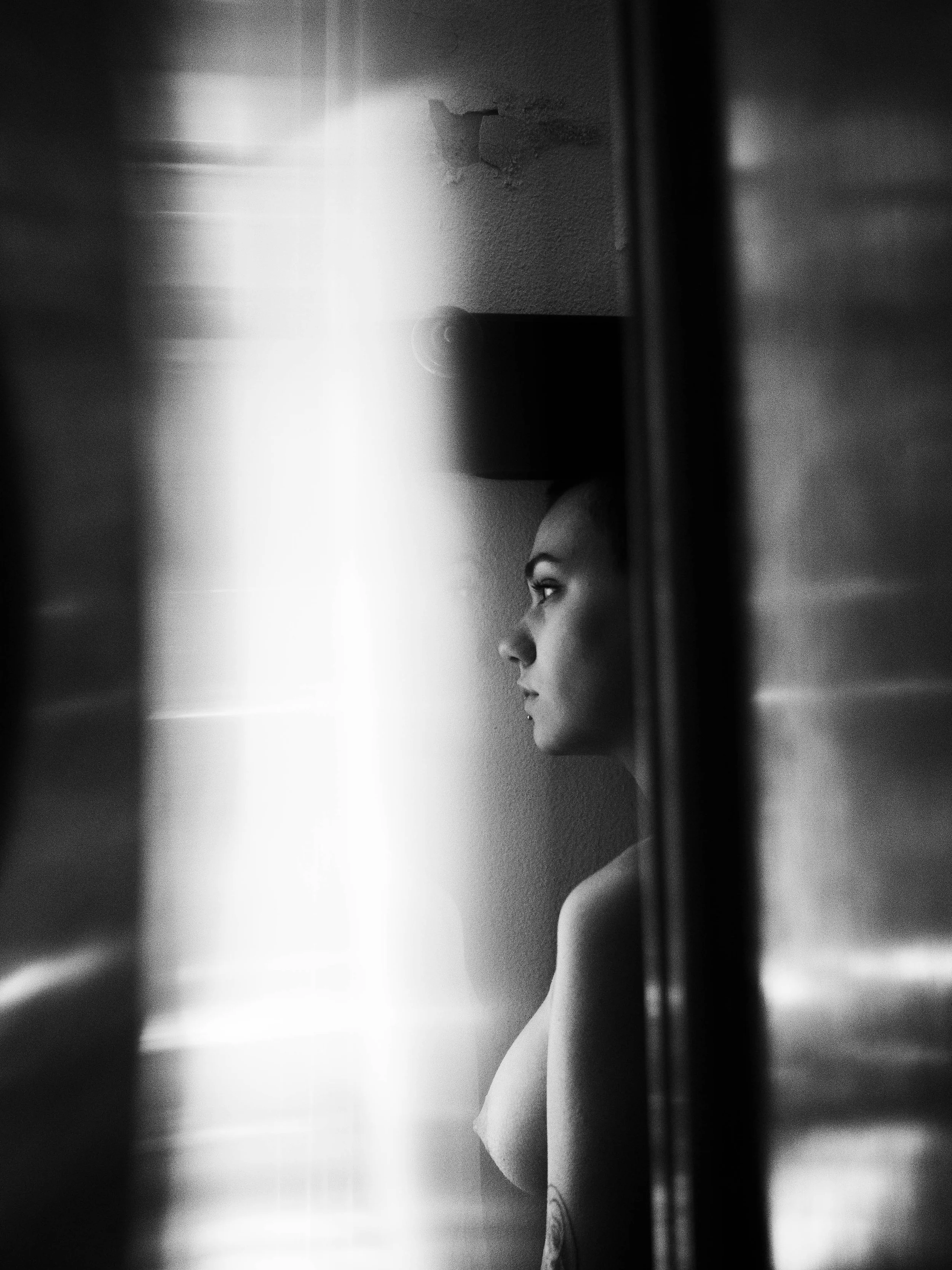 Black and white photo of a woman seen through a narrow gap or doorway, with her side profile visible. She has a serious expression and appears to be looking forward.