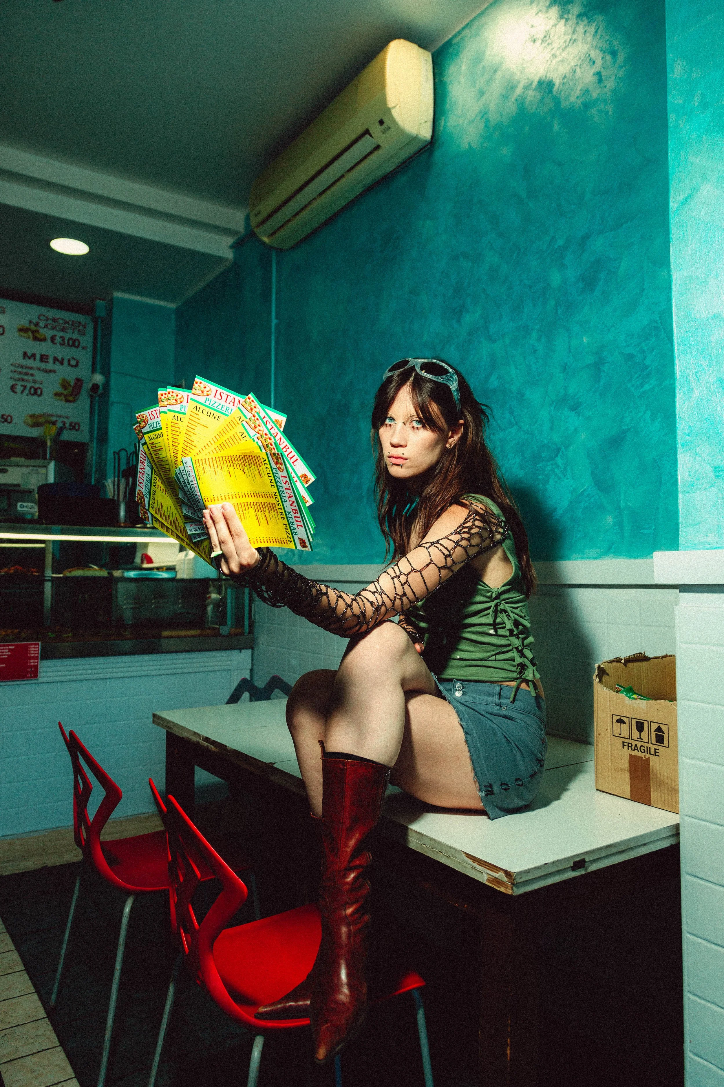 A young woman with long brown hair, wearing a green shirt, shorts, and tall red boots, sitting on a counter in a fast food restaurant, holding multiple yellow menus.