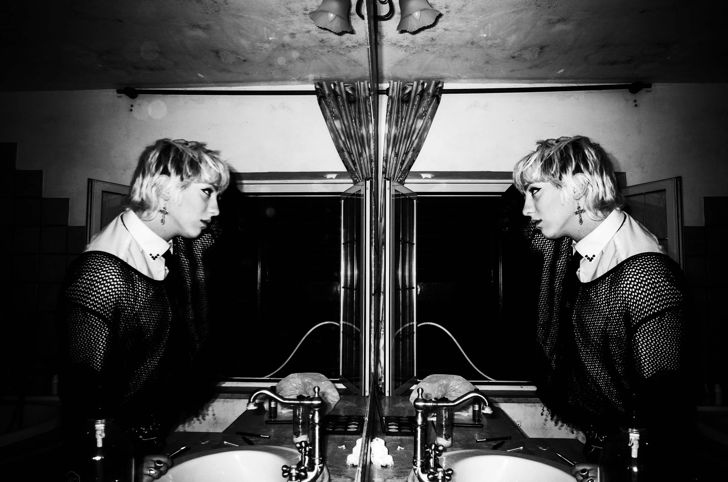 A black and white photograph of a woman looking into a mirror, showing the same woman on both sides of the reflection. She has short, wavy hair and is wearing a mesh top with a white collared shirt underneath and earrings. The setting appears to be a