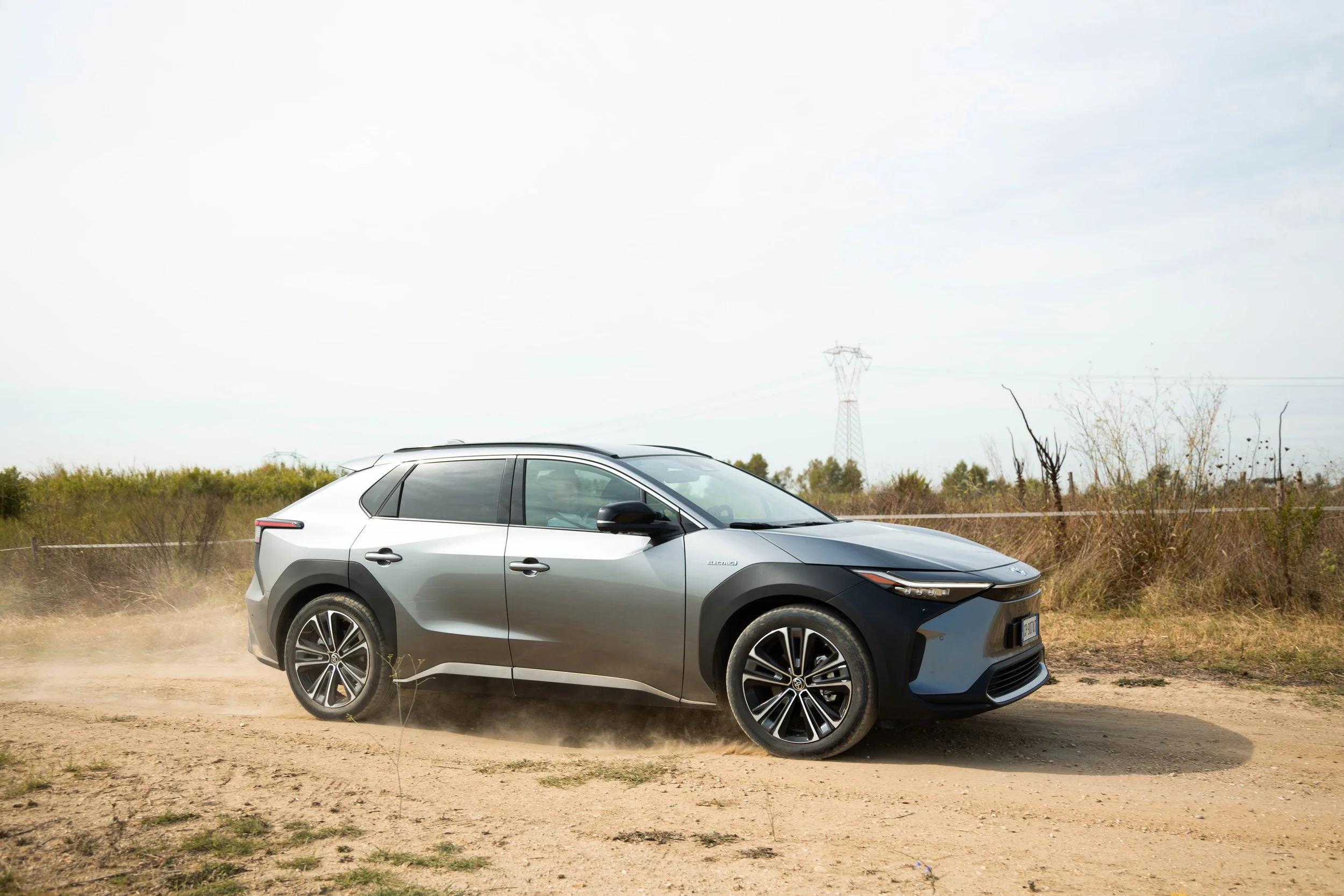 A silver and black electric SUV driving on a dirt road in a rural area with sparse vegetation, power lines in the background, and a partly cloudy sky.