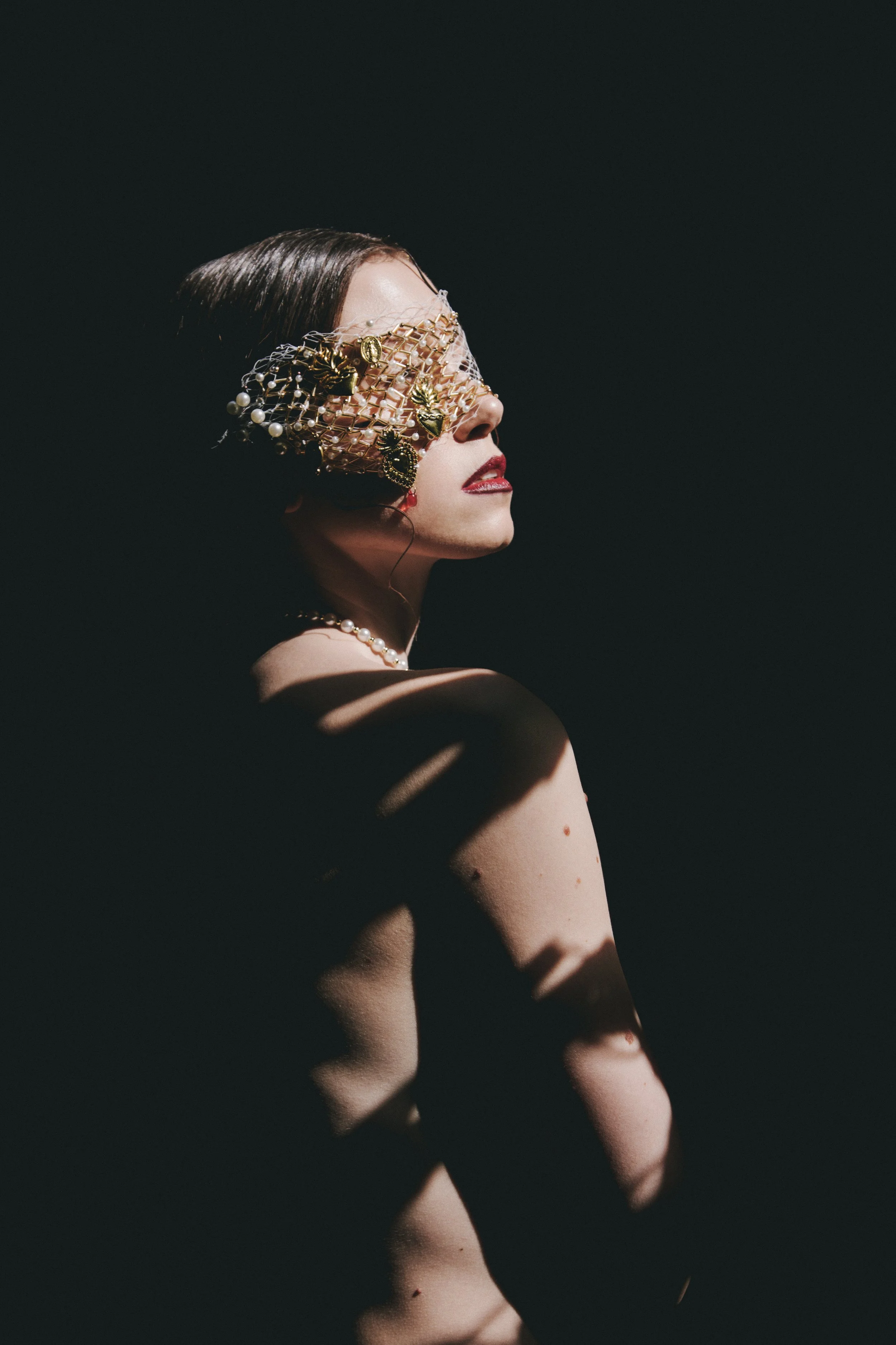 A woman with dark hair and red lipstick wearing an ornate eye mask and pearls, with dramatic shadow play on her shoulder and arm.