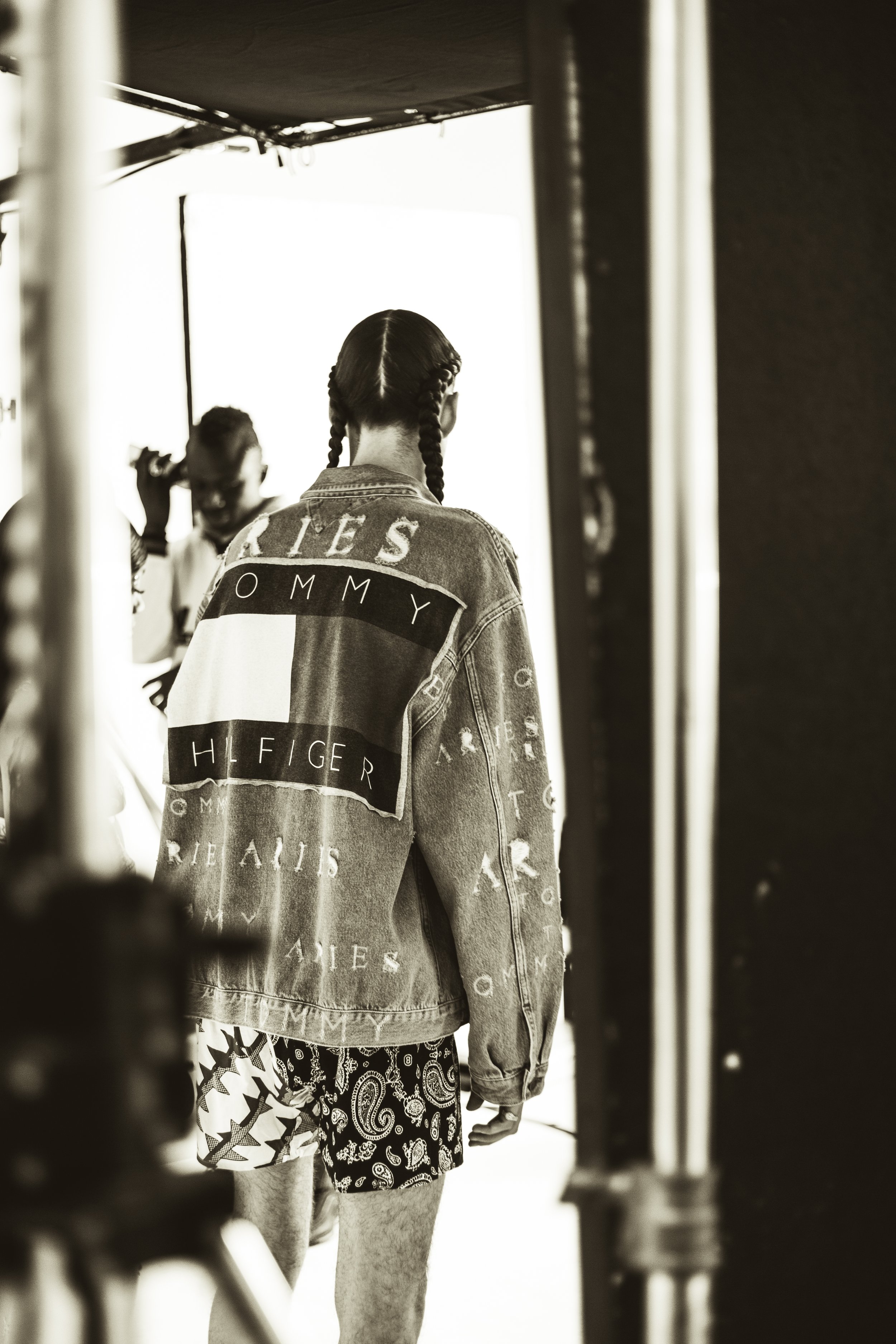 Behind-the-scenes of a photoshoot showing a woman in a denim jacket with Tommy Hilfiger logo and patterned shorts, with crew members in the background.