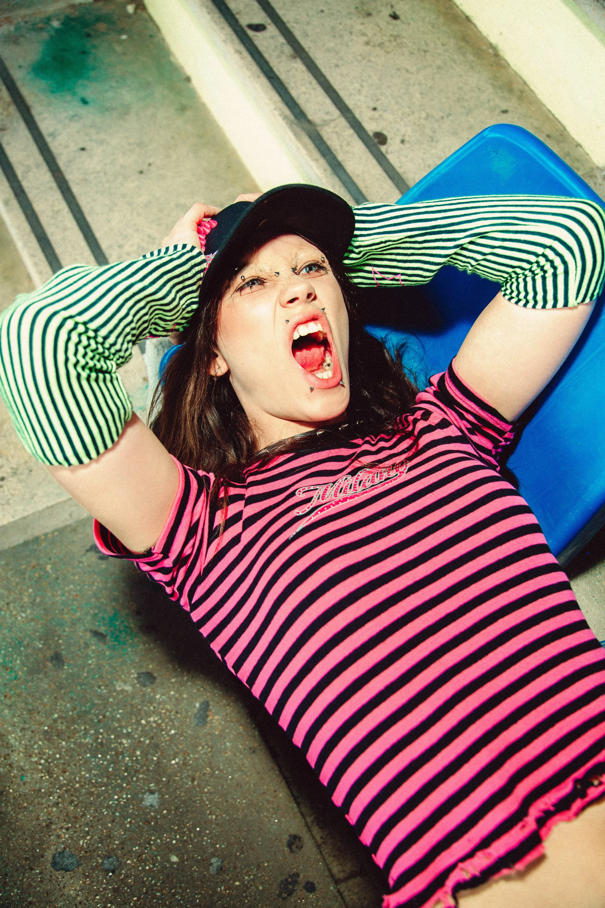 A woman lying on the ground with her hands on her hat, wearing a pink and black striped shirt and green and black striped arm warmers, appears to be screaming or shouting.