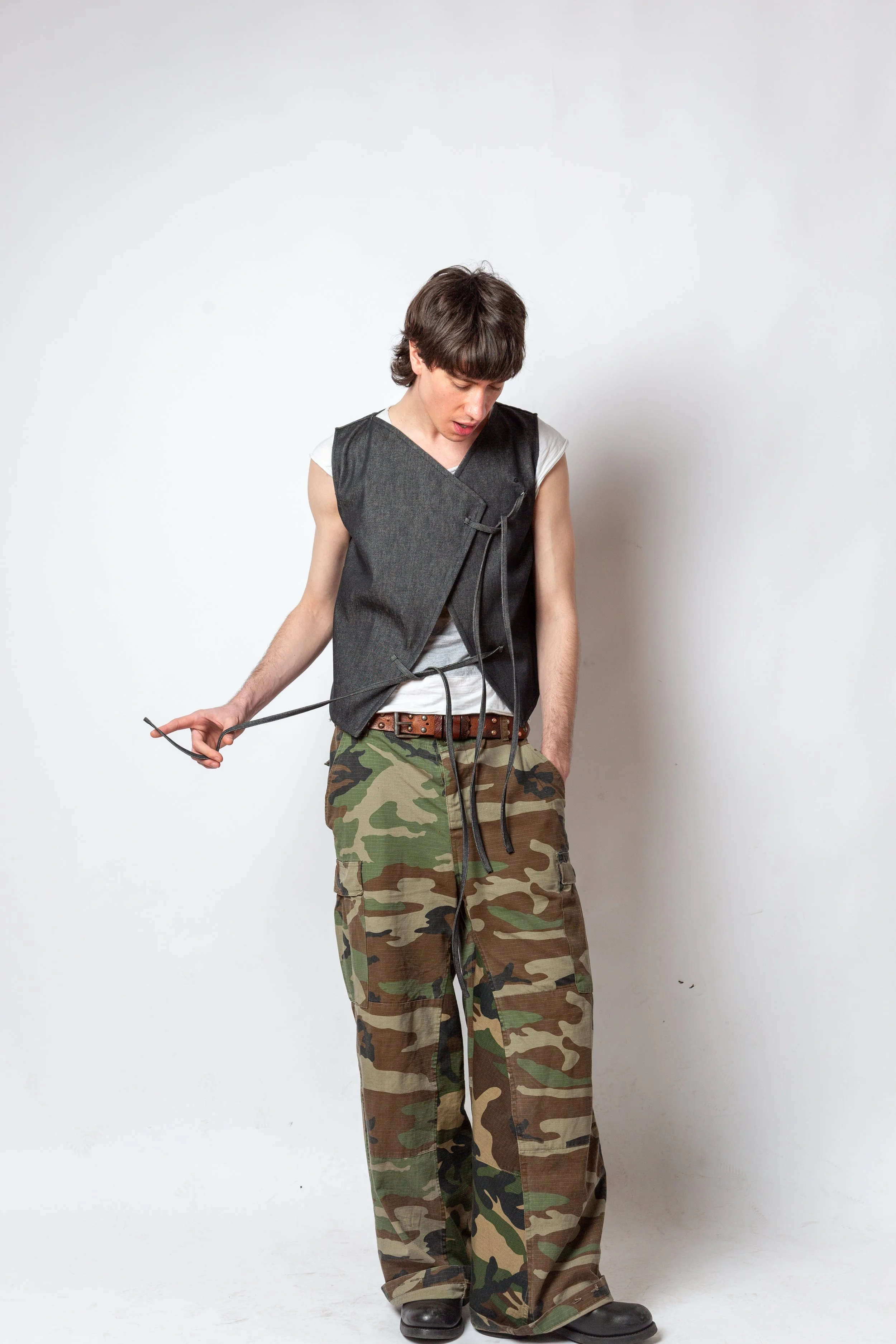 Young man with dark hair wearing camouflage pants, a gray vest, and a white t-shirt, looking down and holding a black cord against a plain white background.