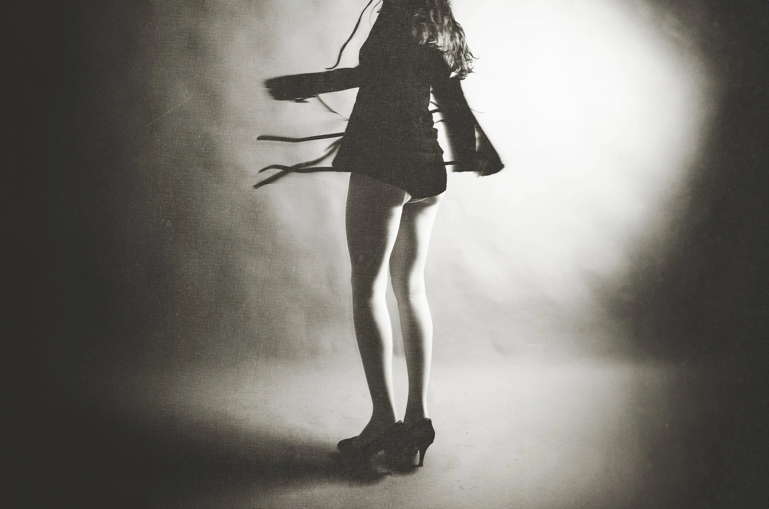 Black and white photo of a woman from behind, wearing a short dress and high heels, with long hair and ribbon-like accessories flowing around her.