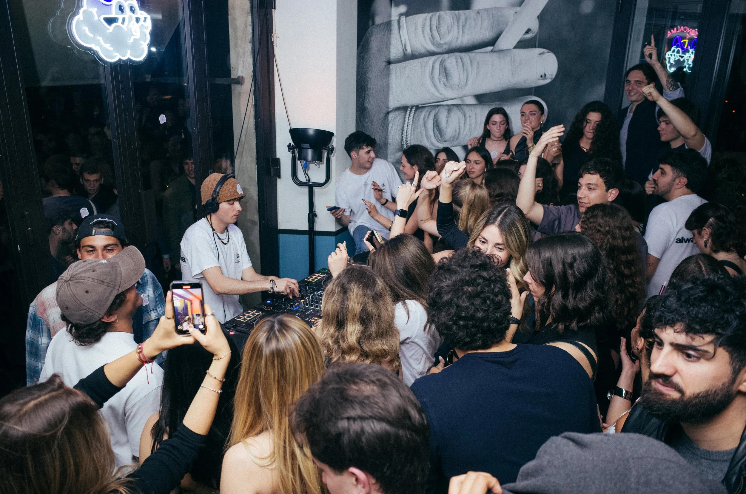 Crowded indoor party with a DJ playing music, people dancing, taking photos, and enjoying themselves.
