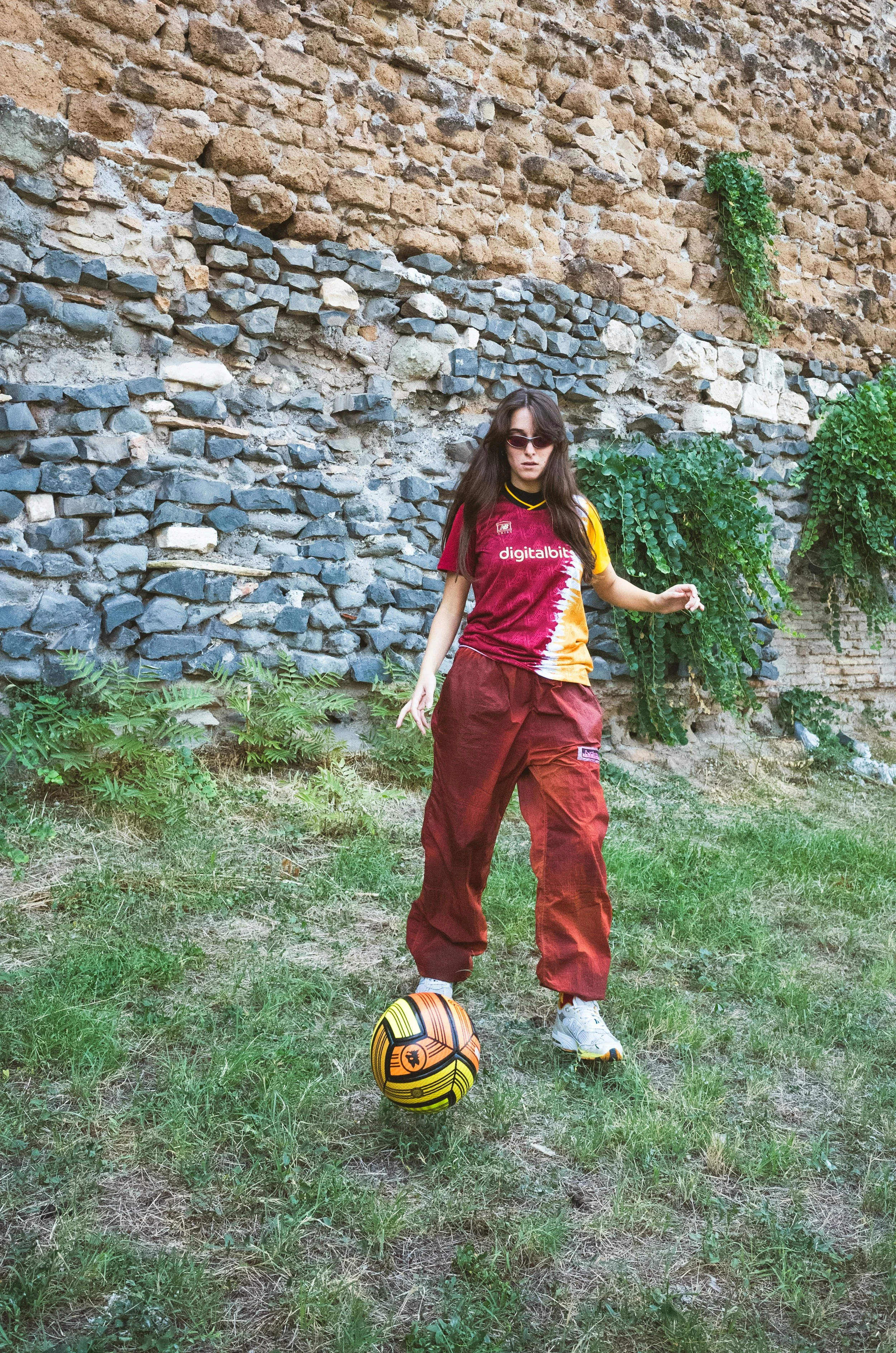 A young woman wearing sunglasses, a red and yellow sports jersey, and red baggy pants is playing with a soccer ball outdoors on a grassy area with a stone wall and green plants in the background.