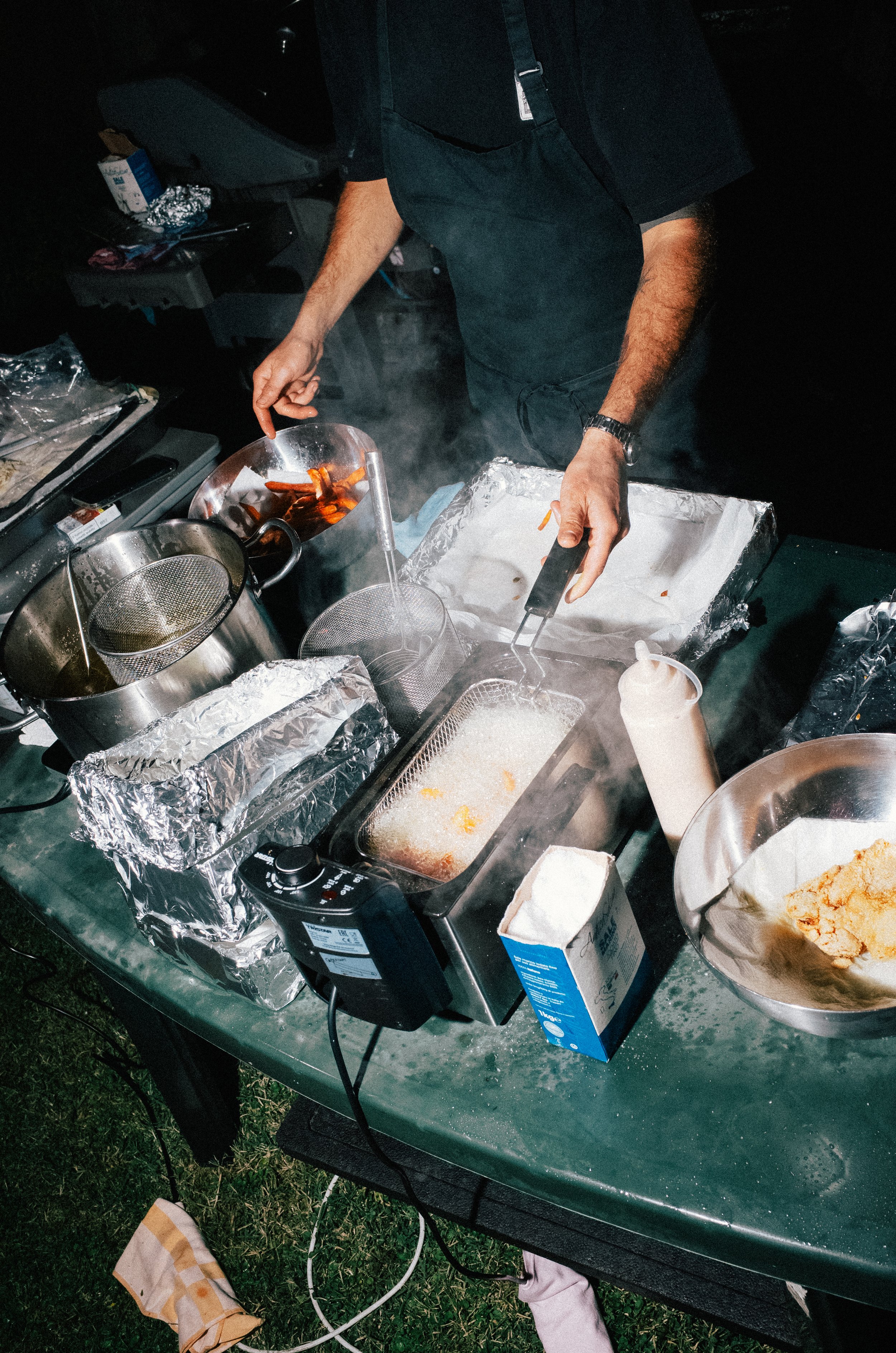 Person cooking food on a green outdoor table at night, with open foil-wrapped containers, a box of milk, a bowl of fried food, and cooking utensils.