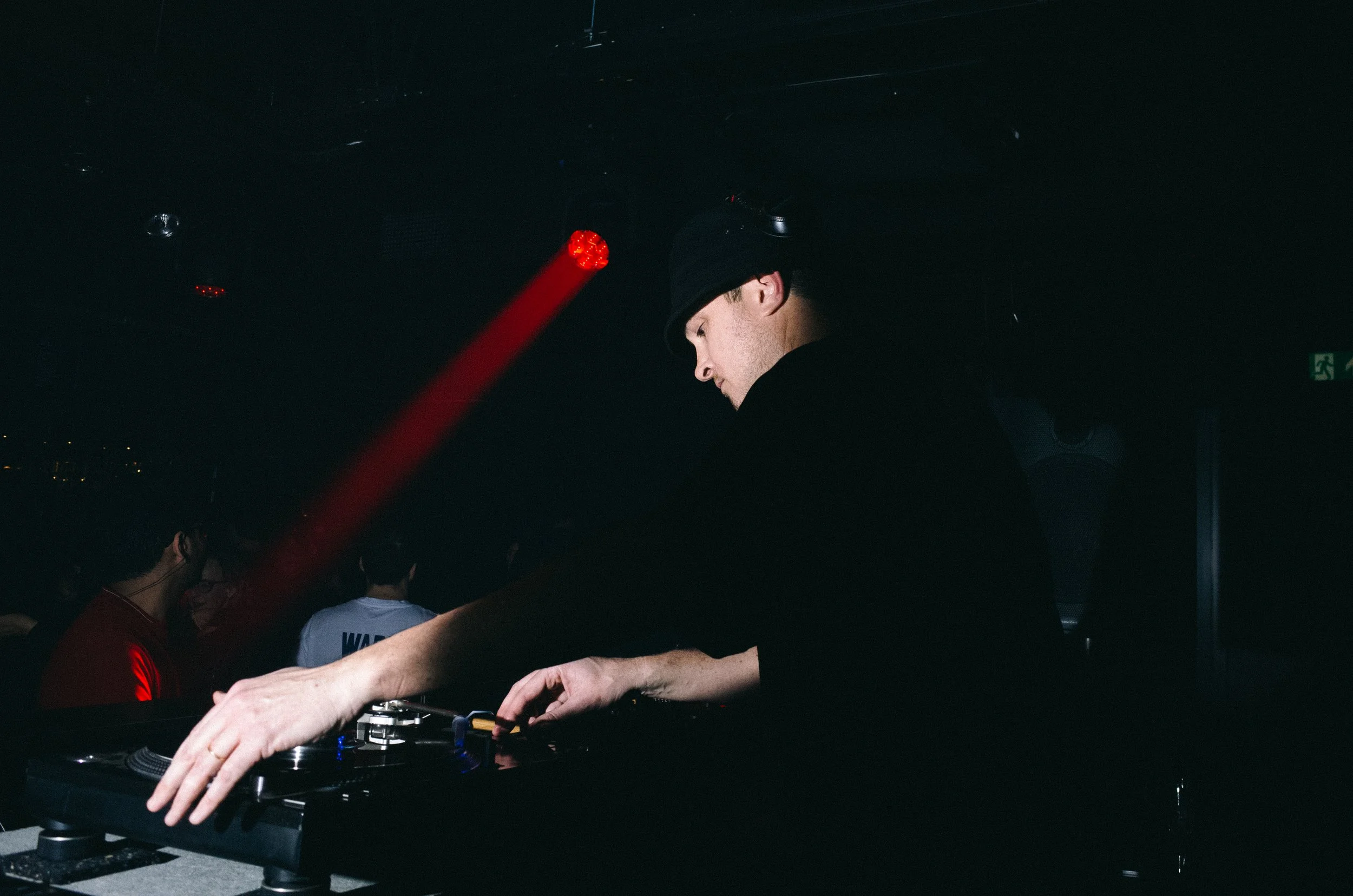 A DJ wearing a black hat and black shirt, operating a turntable in a dark nightclub with red and white lighting and a crowd in the background.