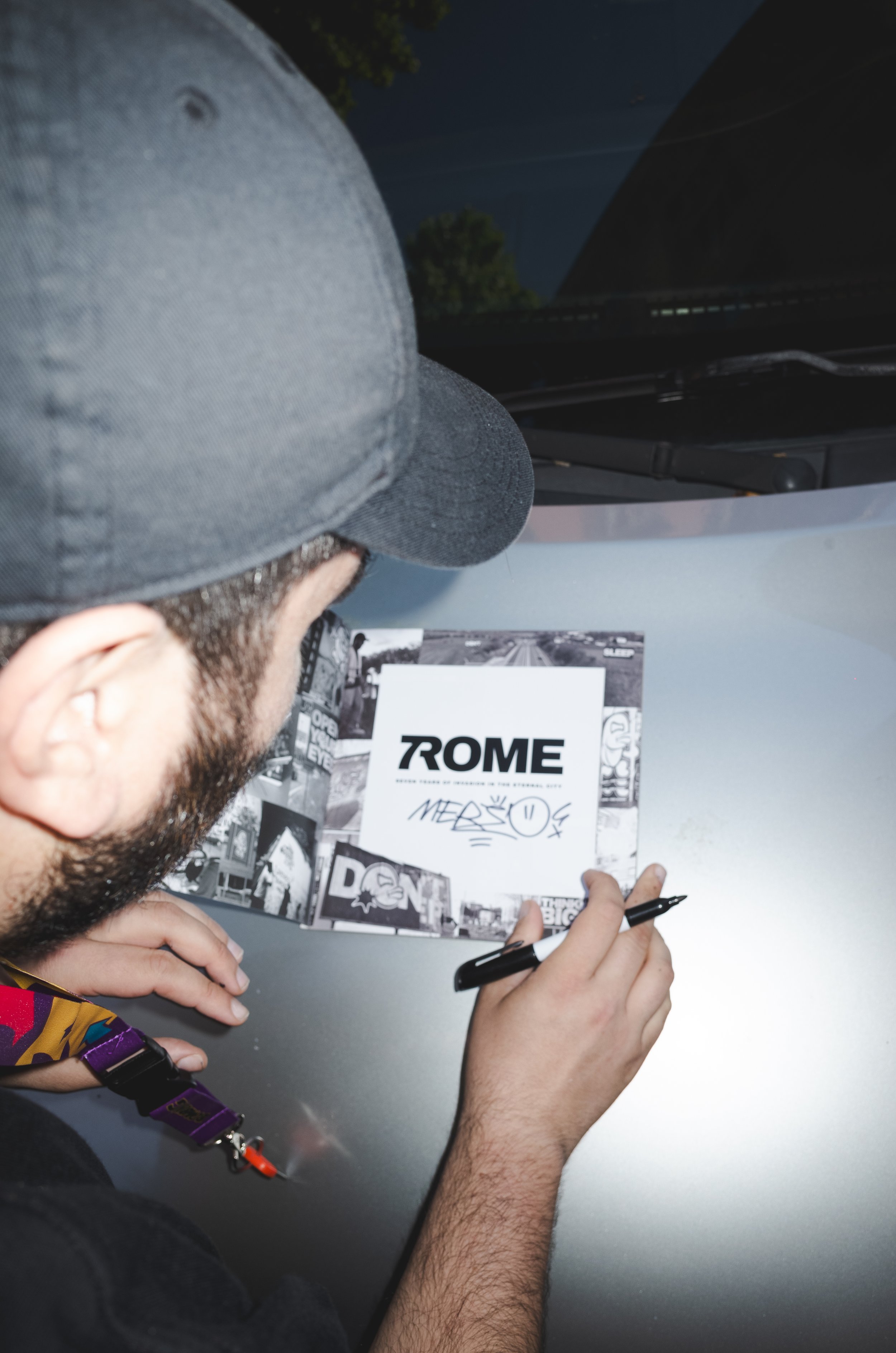 A person with a beard and cap signing a poster that features the word "TROME" and a black-and-white photo collage, while a car and some trees are seen in the background.