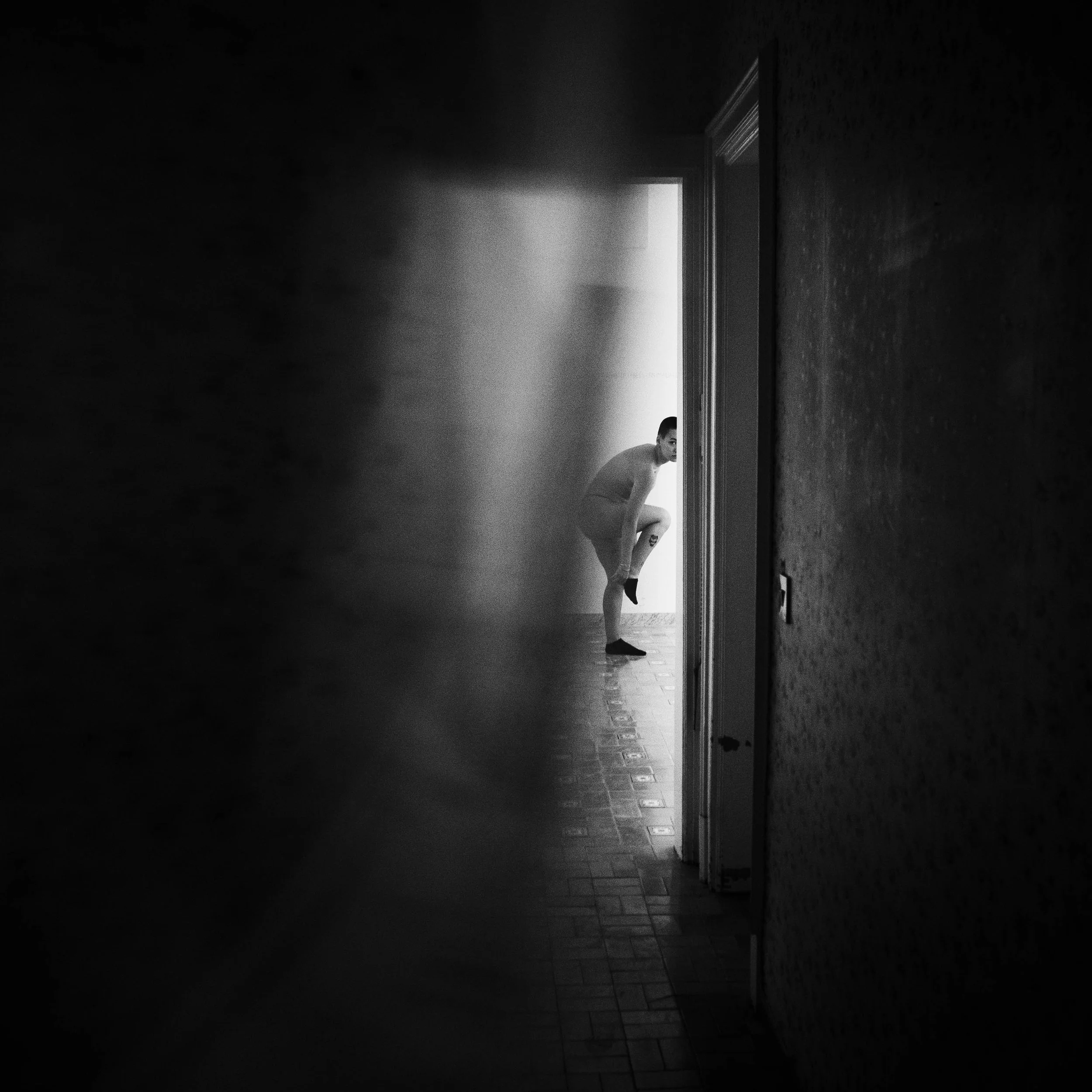 Black and white photo of a person seen through a slightly open door, bending down and adjusting their sock in a dimly lit hallway