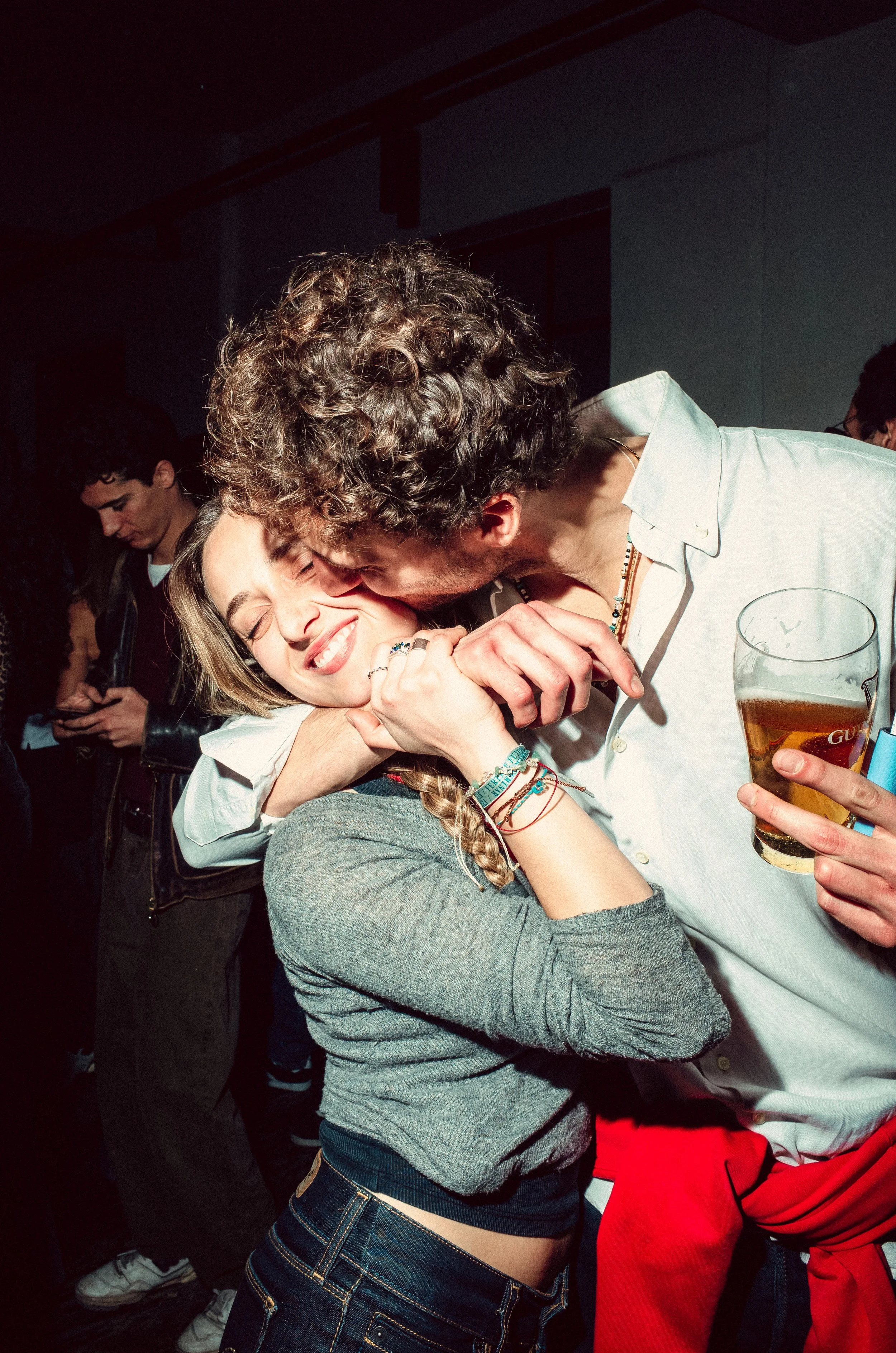 A man kissing a woman on the cheek while she smiling with her eyes closed at a party, holding a beer glass.