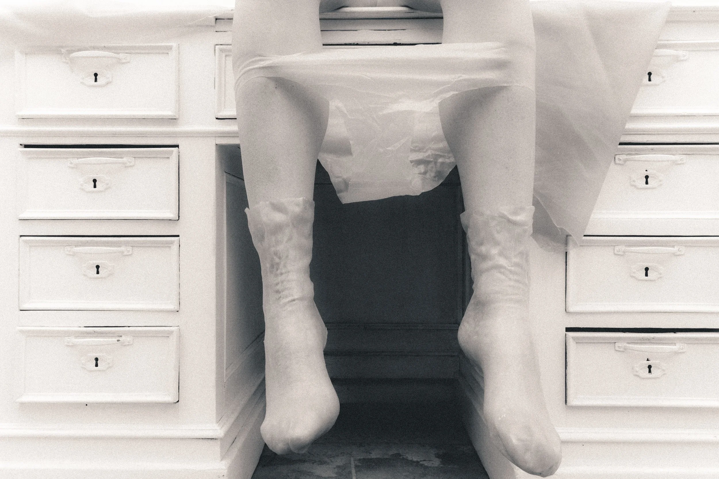 A person sitting at a desk with their legs hanging down, wearing white socks. The desk has multiple drawers with small keyholes, and the photo is taken from underneath, focusing on the person's legs and the desk.