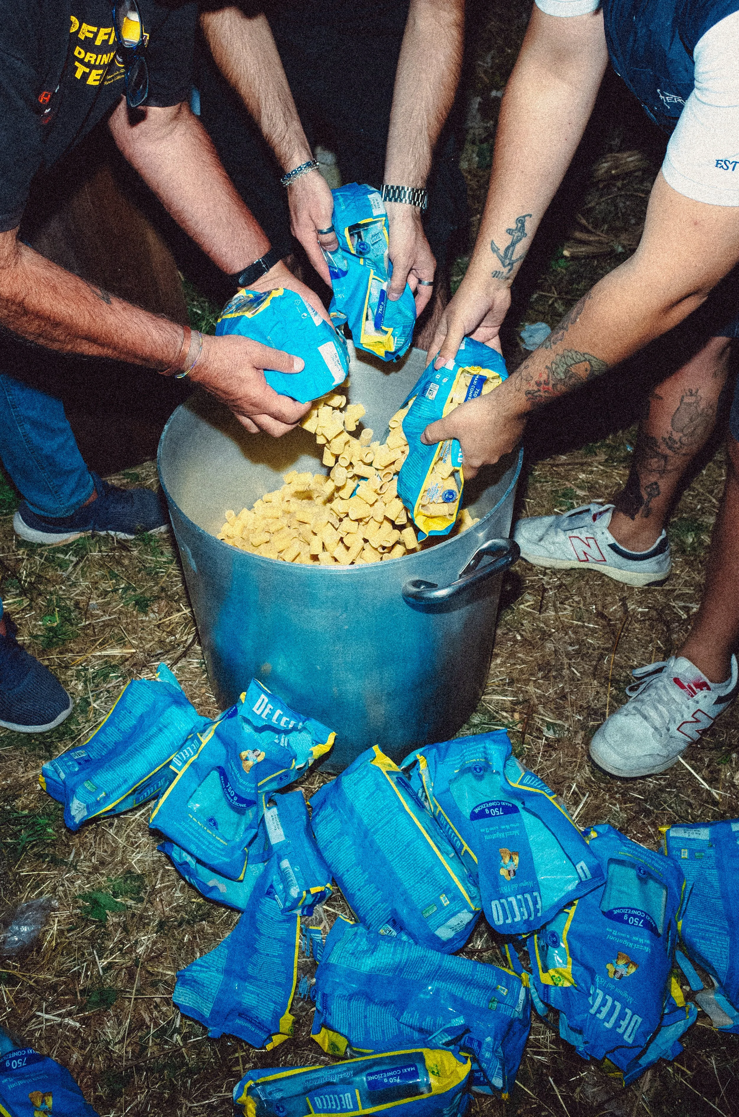Multiple people gather around a large pot of pasta outdoors, pouring it into the pot from blue packaged bags.