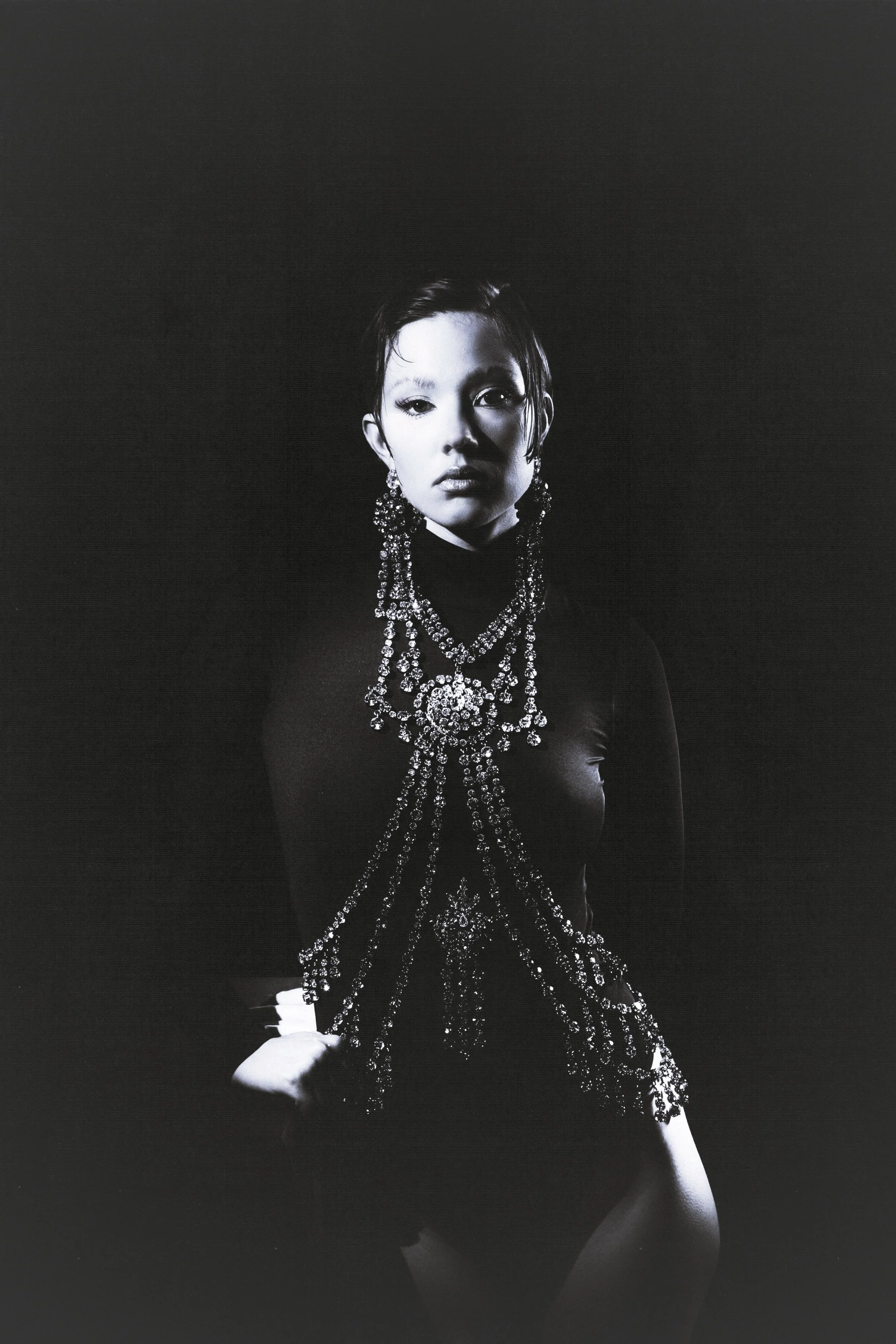 Black and white portrait of a woman wearing elaborate jewelry and a dark, fitted outfit, standing against a dark background.