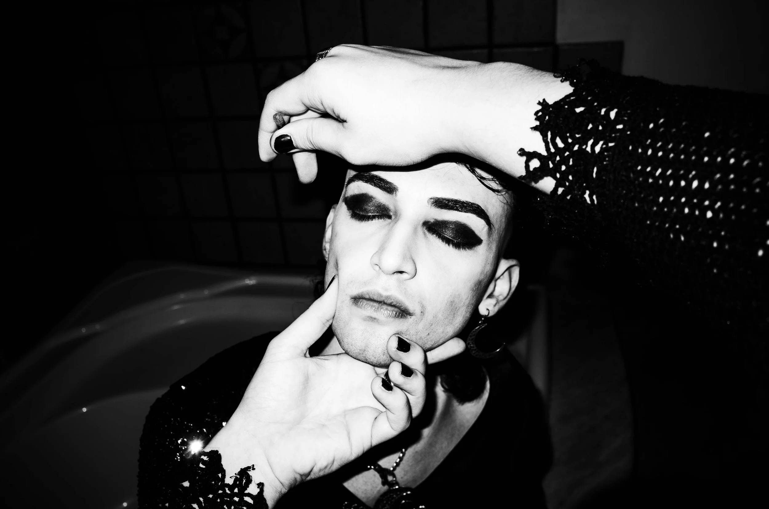 A black-and-white photo of a person with makeup, eyes closed, receiving a facial massage with their hands.