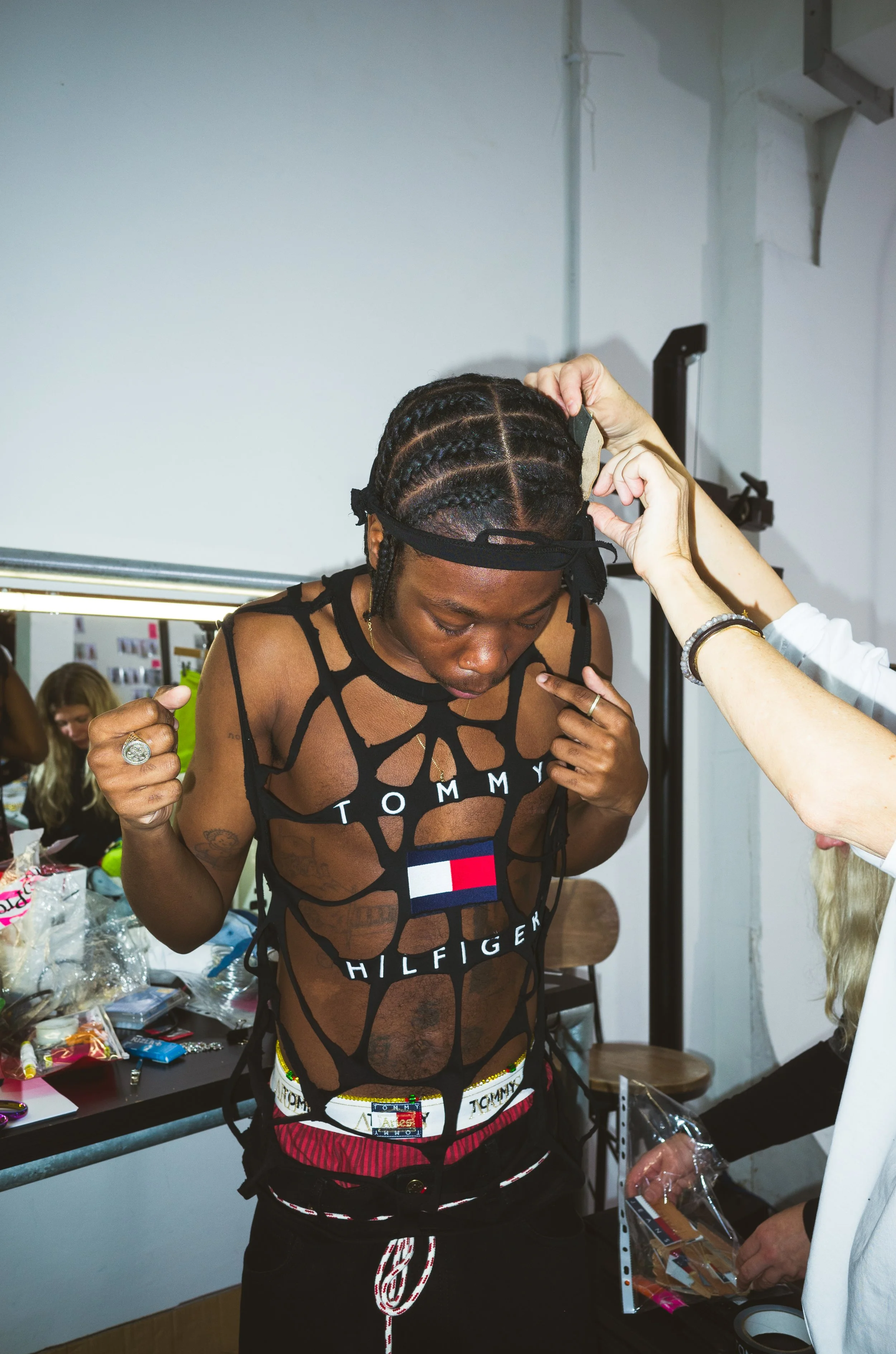 A person with braided hair and a black Tommy Hilfiger tank top is getting fitted with a wig or headpiece by a stylist in a room with various items on a table.