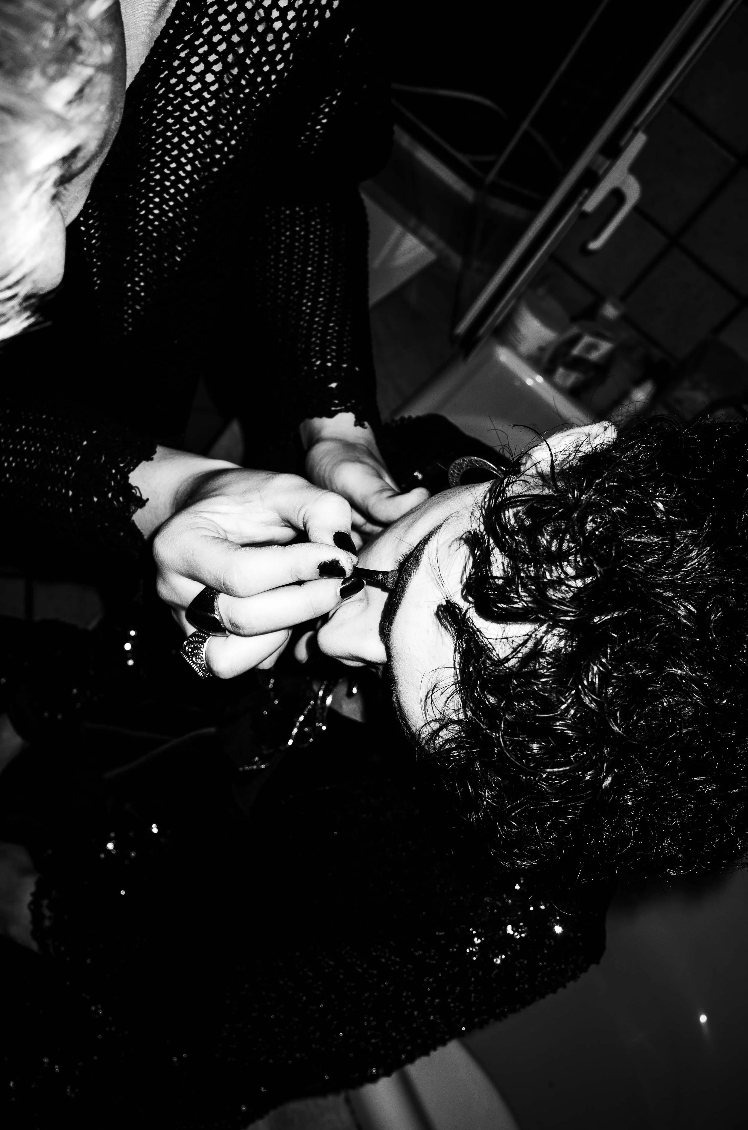 A person with curly hair getting their makeup applied by another individual wearing a black, perforated dress in a room with cabinets and various items in the background.