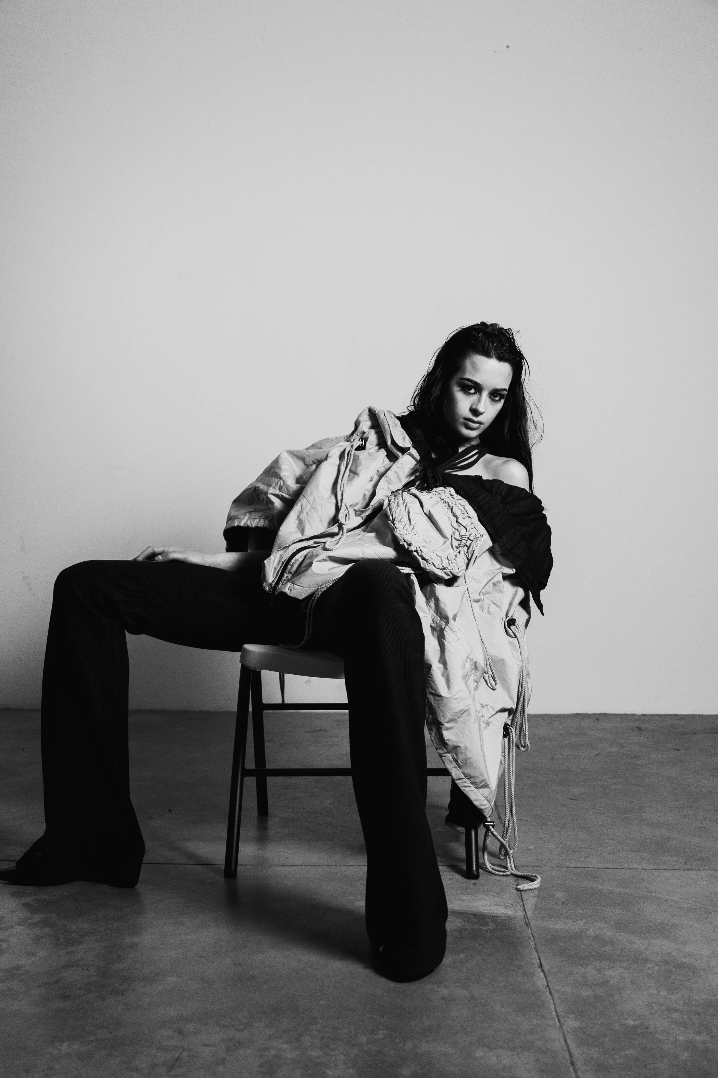 A young woman with dark hair sitting on a chair against a plain wall, wearing loose black pants and a large, light-colored jacket, with an intense expression on her face.