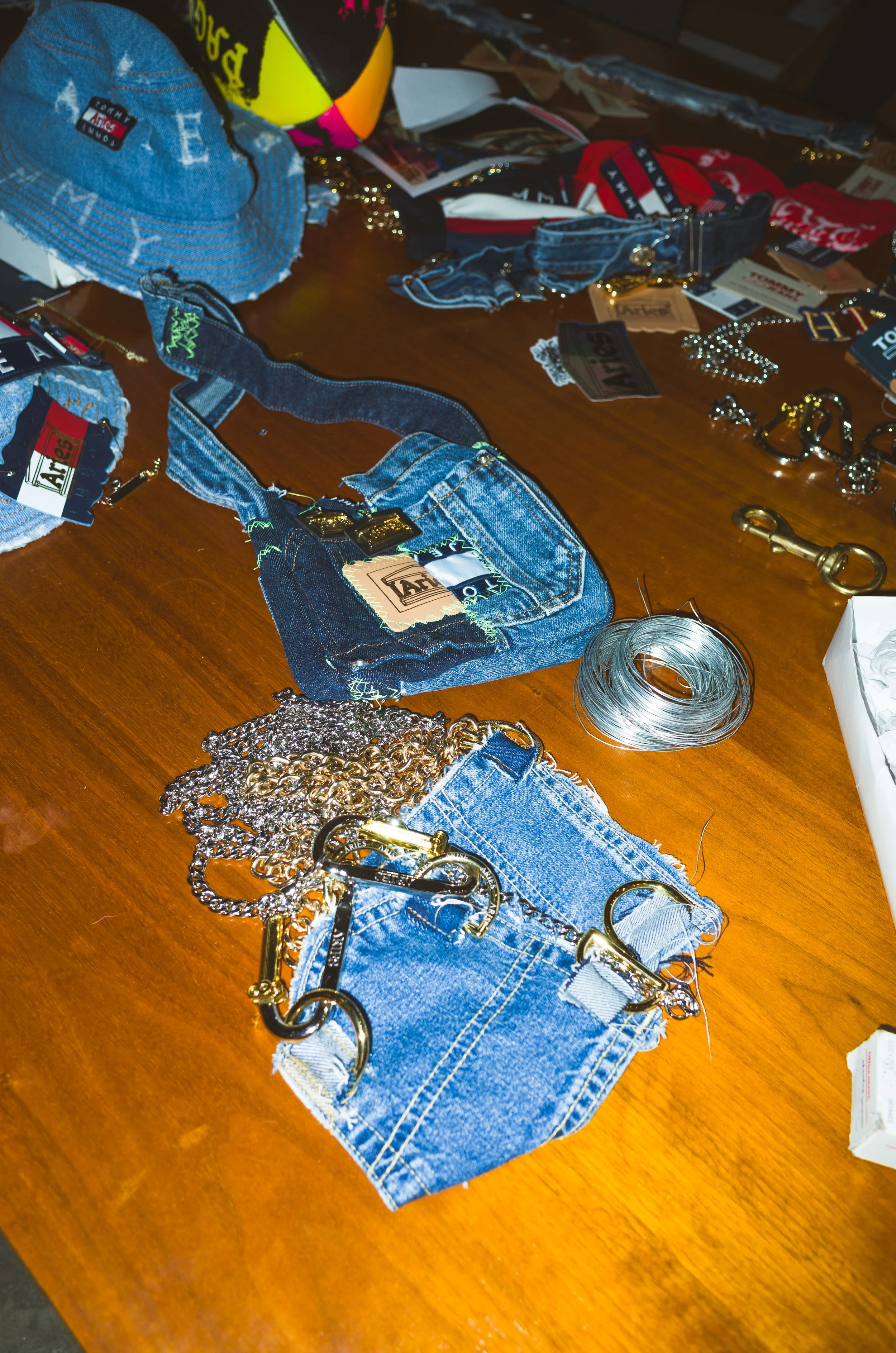 A wooden table covered with various denim clothing items, jewelry, and accessories. There are denim bags, a pair of denim shorts with gold clips, a small denim purse, metallic jewelry, and assorted small items, including tags and keys, scattered acro