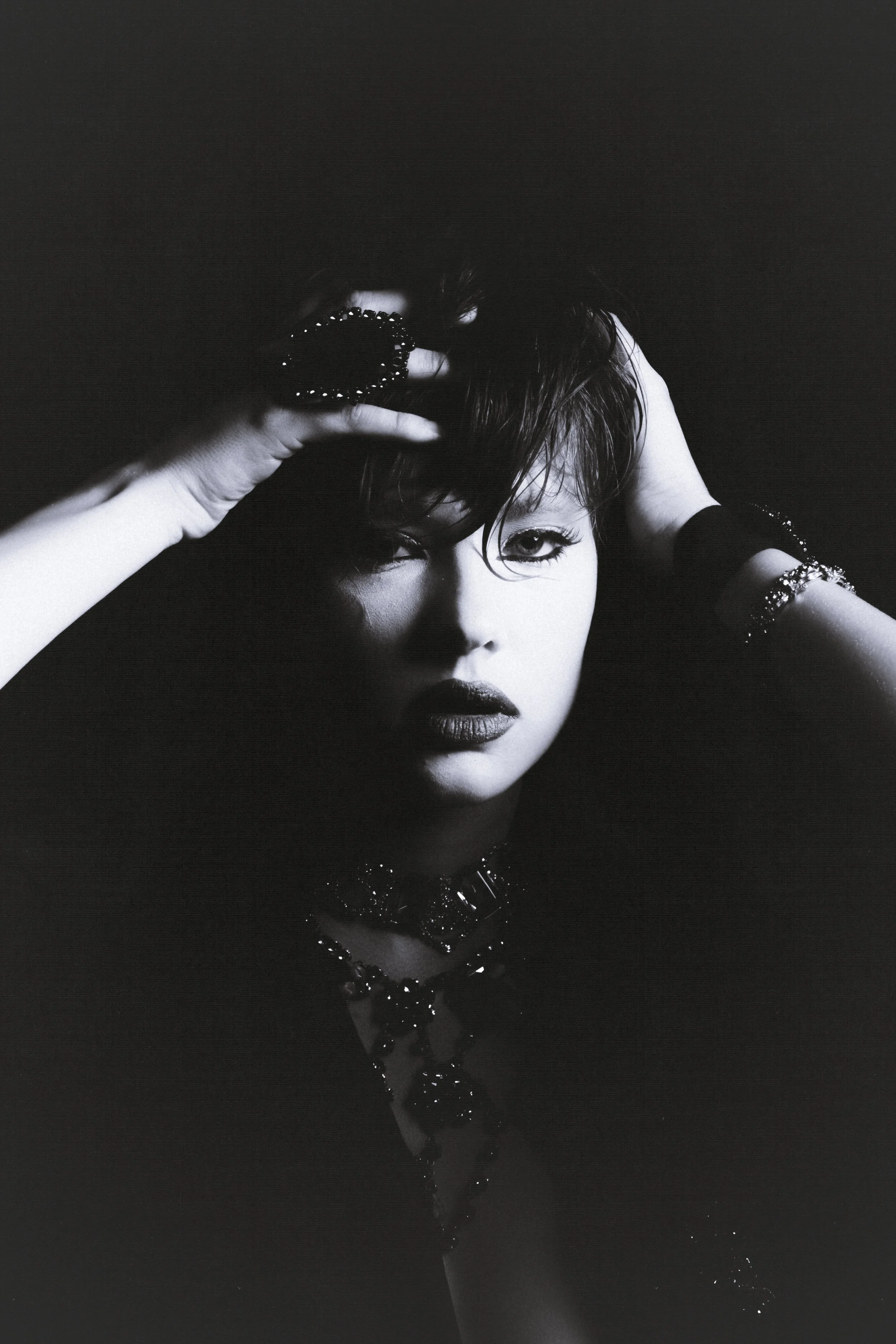 Black and white portrait of a woman posing with one hand in her hair, wearing dark lipstick, makeup, and jewelry.