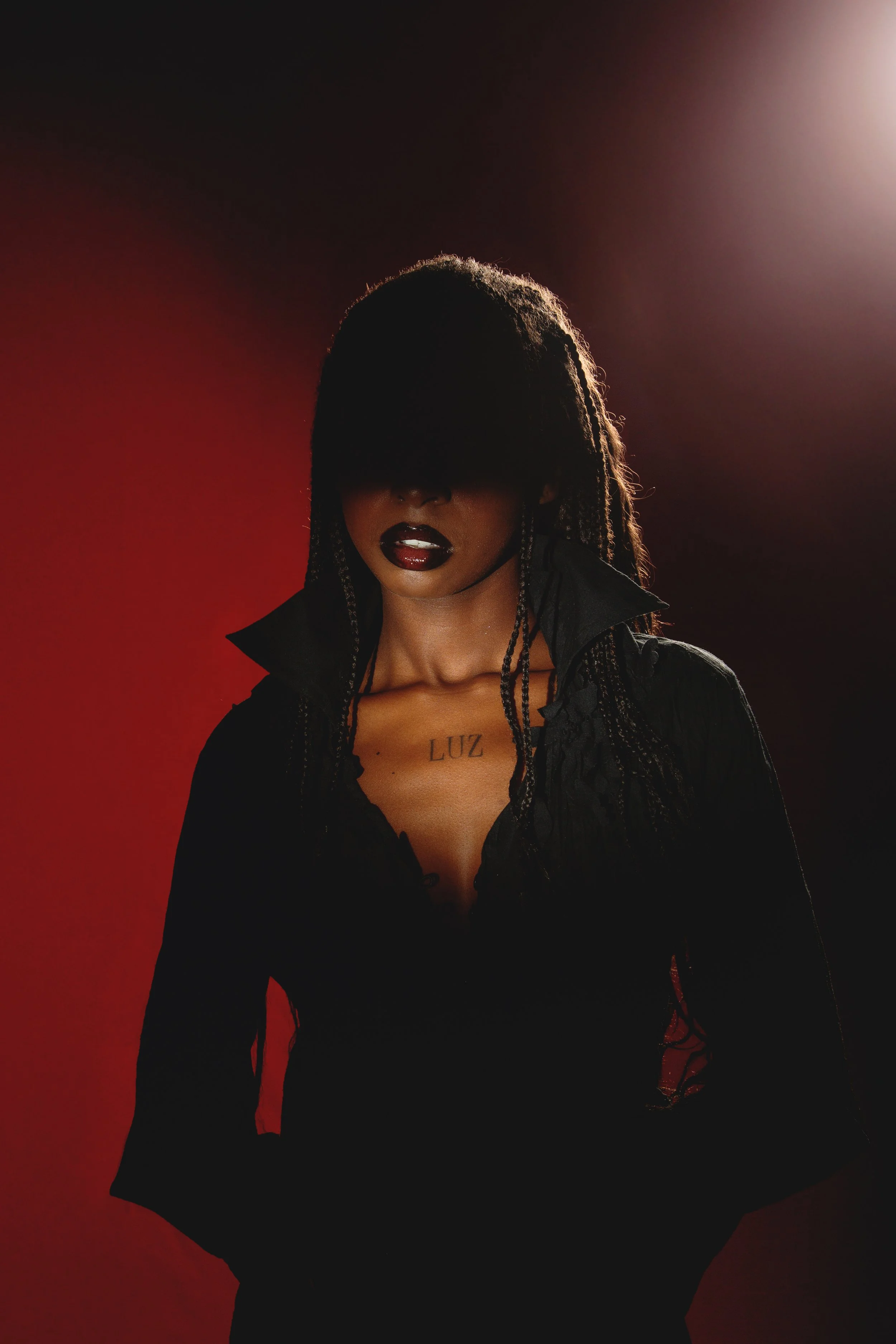 A woman with dark braided hair, wearing dark lipstick and a black jacket, stands against a dark red background with bright lighting from behind, casting her face in shadow. A tattoo on her chest reads "LUZ".