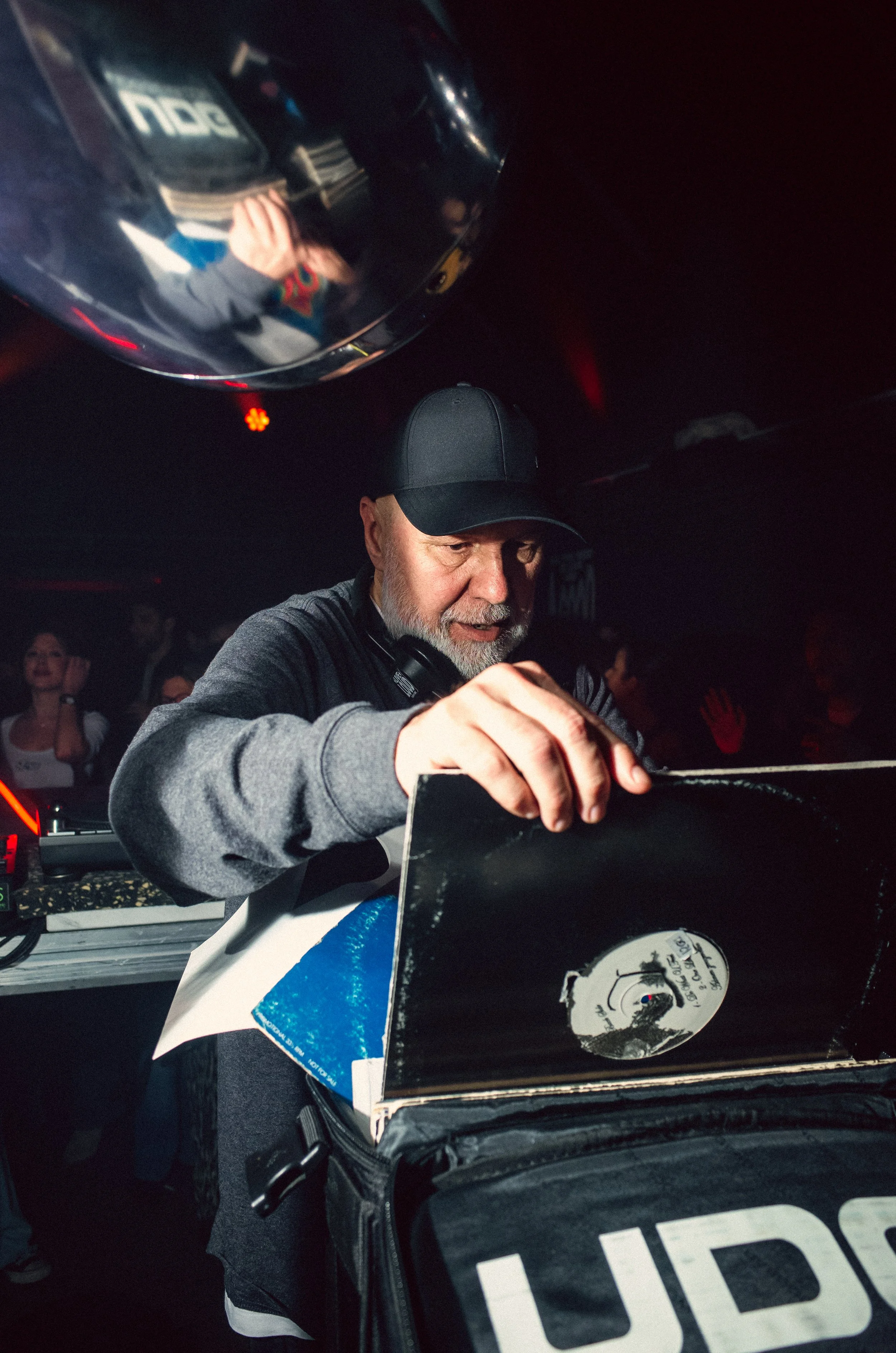 A DJ with a gray beard and black cap playing music at a nightclub, surrounded by people, with a reflective disco ball overhead.