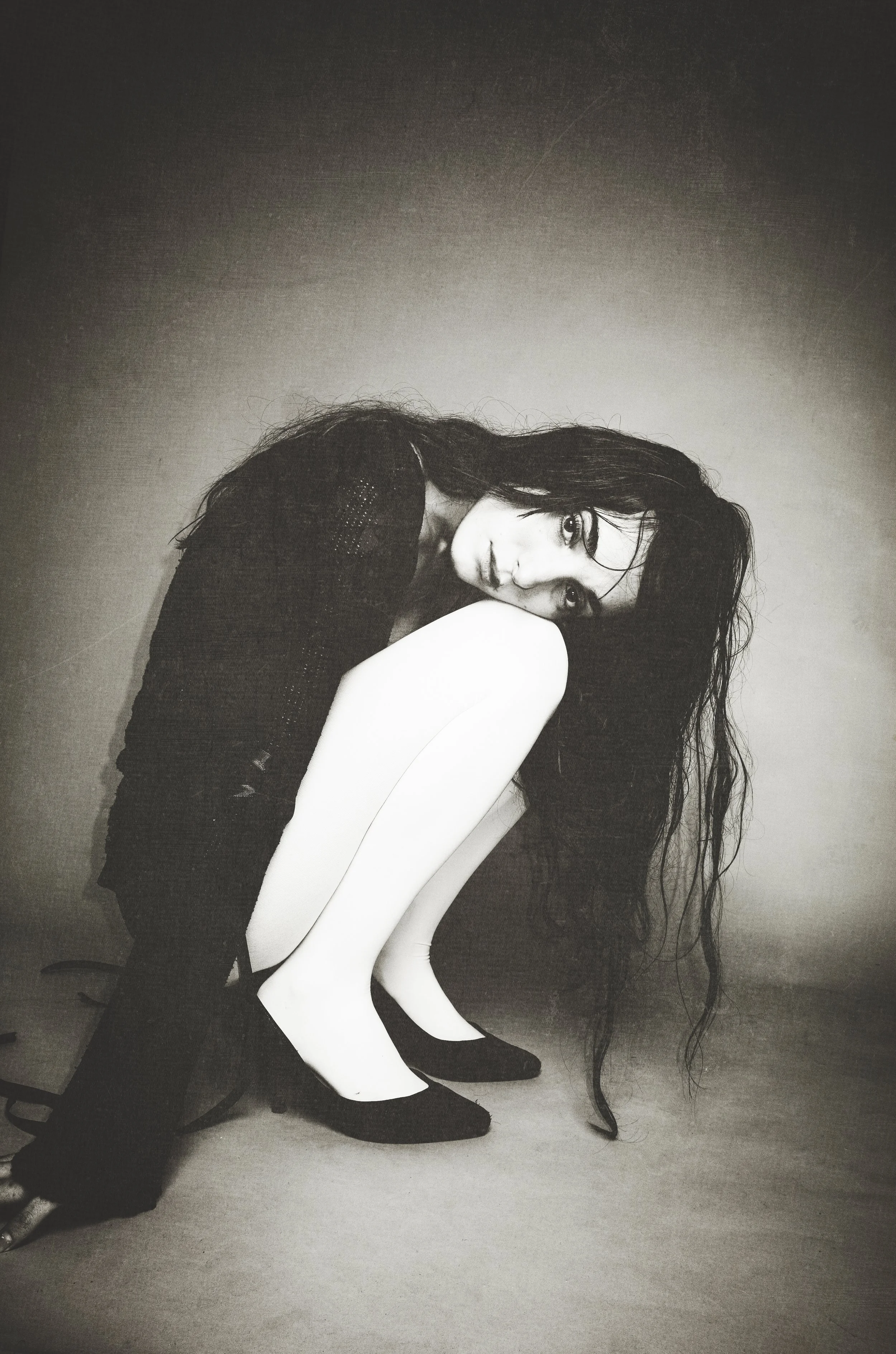 Black-and-white photo of a woman with long, wet hair sitting on the floor with her knees drawn to her chest, looking at the camera, against a plain wall background.