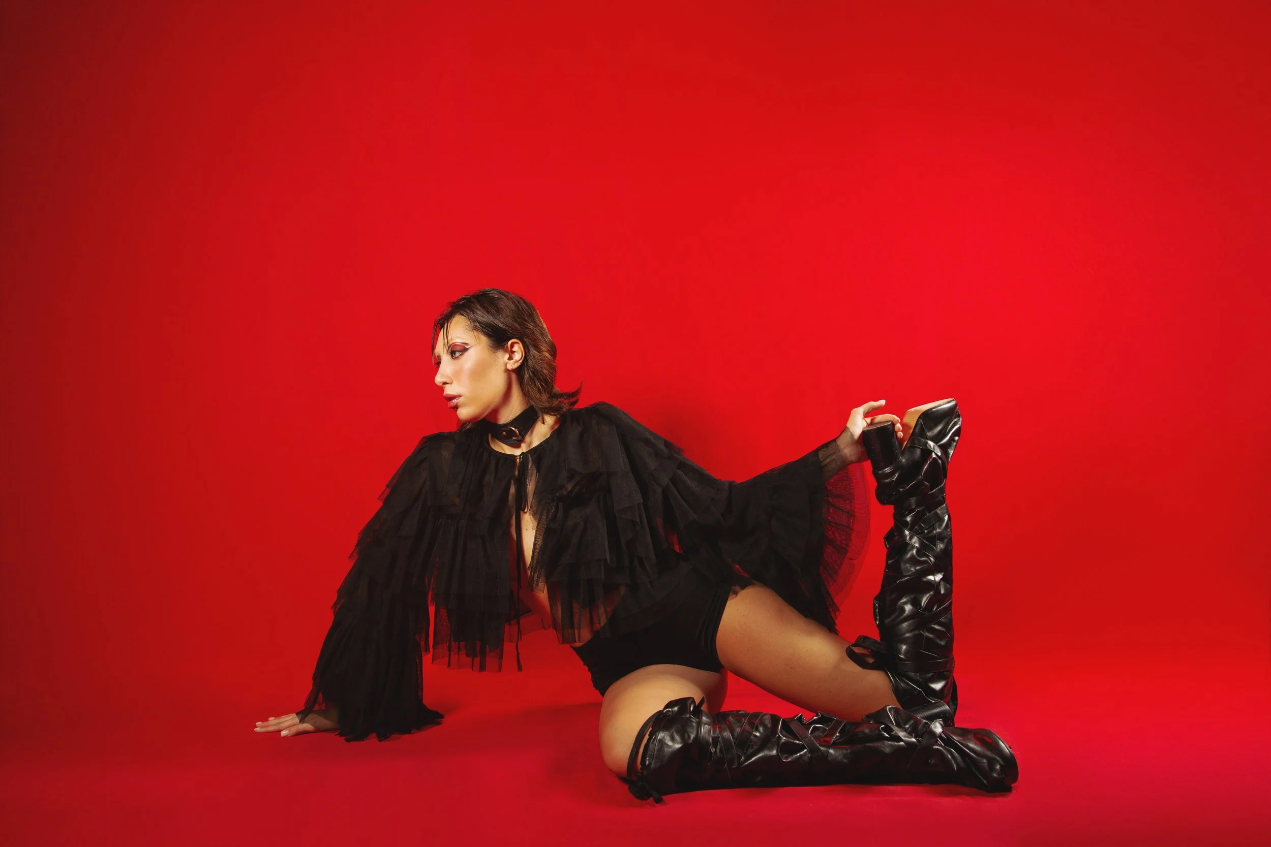 A woman with dark hair in stylish makeup and black clothing, sitting on the floor, stretching her leg while holding her boot against a bright red background.