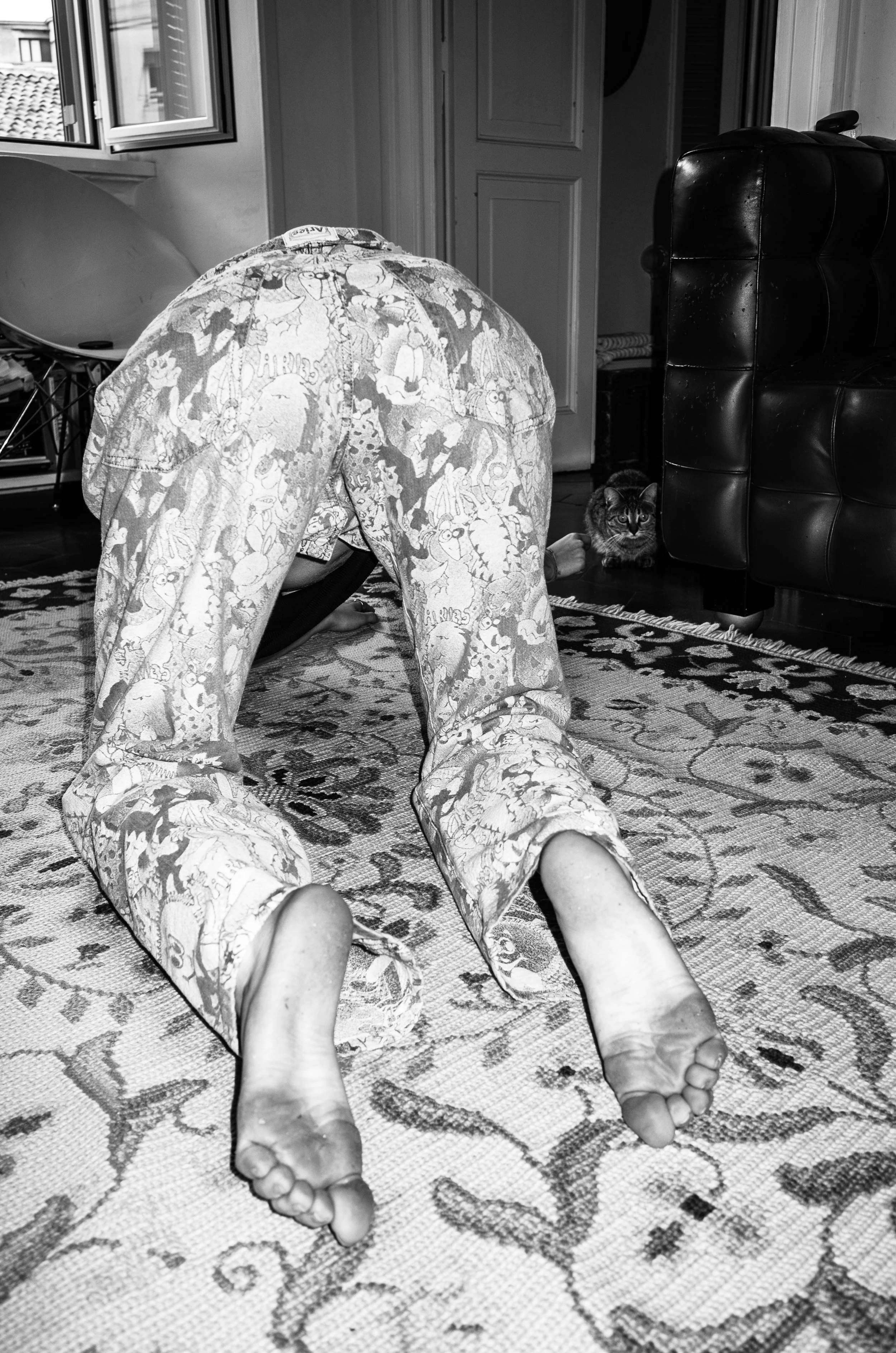 Person in pajamas bending over on the floor with feet visible, and a cat peeking out from behind a piece of furniture, inside a room with a patterned rug, open window, and closet doors.