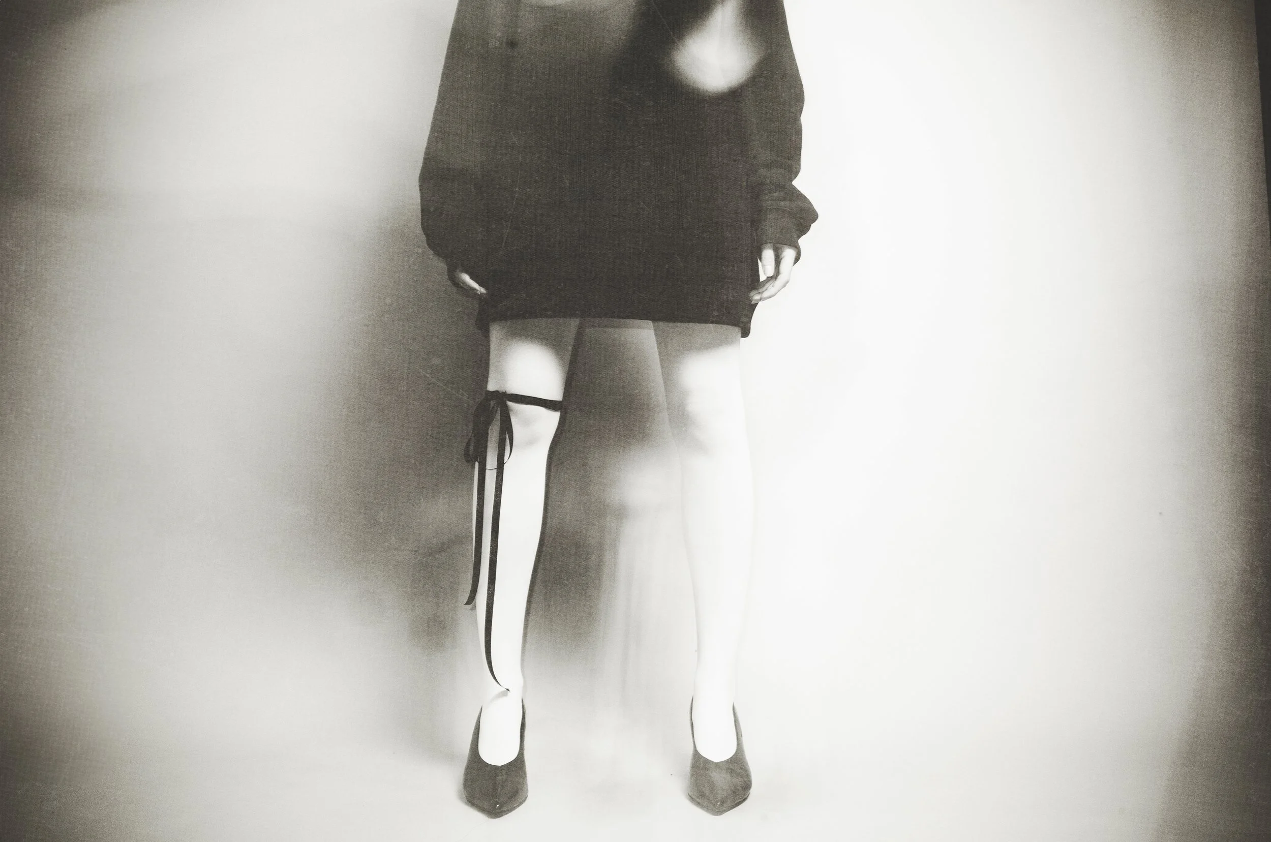 Lower half of a person wearing high heels, a black skirt, a black sweater, and some stockings with a ribbon tied around the left thigh.