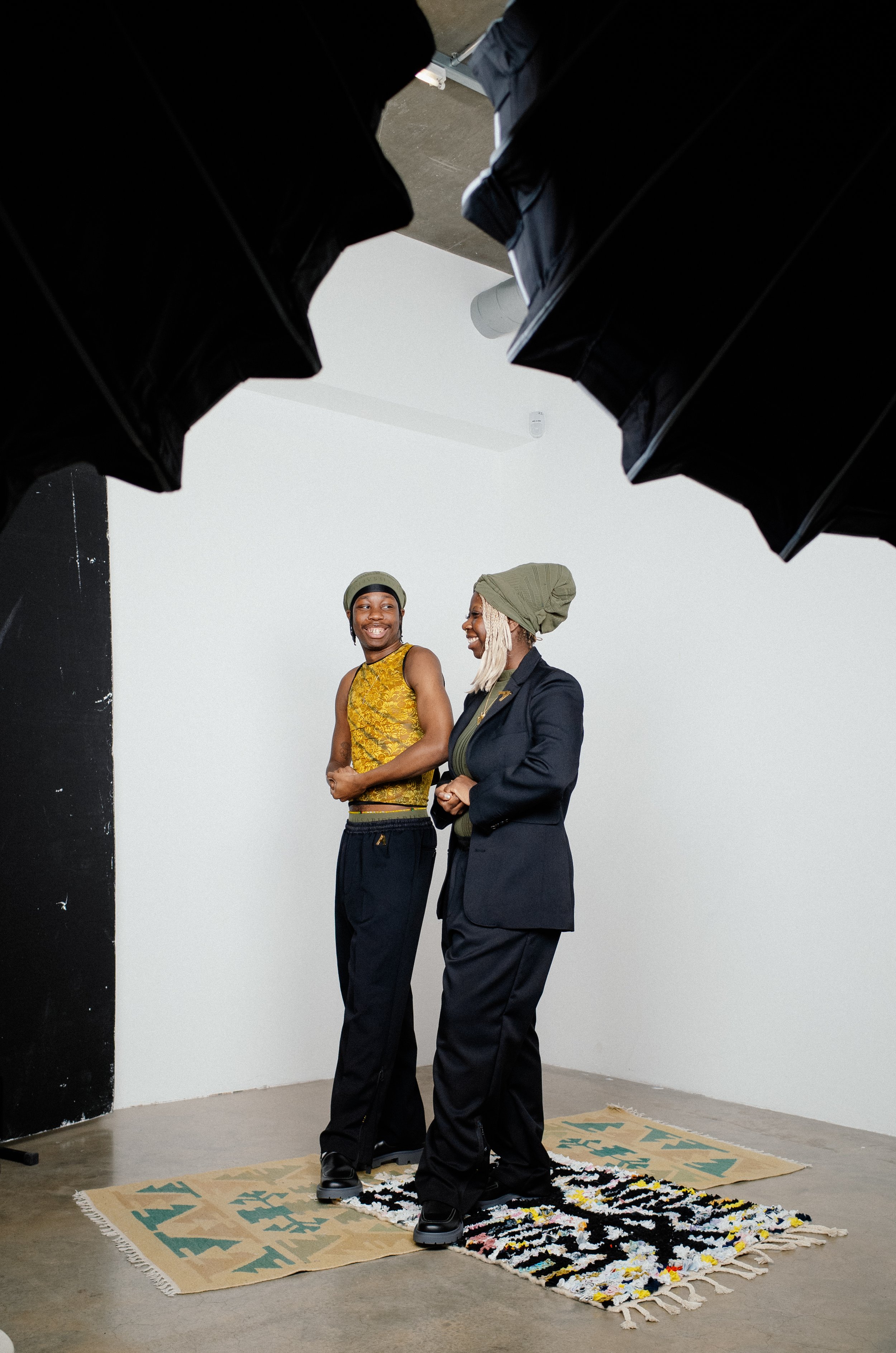 Two women standing on rugs in black suits and head coverings, smiling and talking to each other in a photography studio with black and white backdrops.