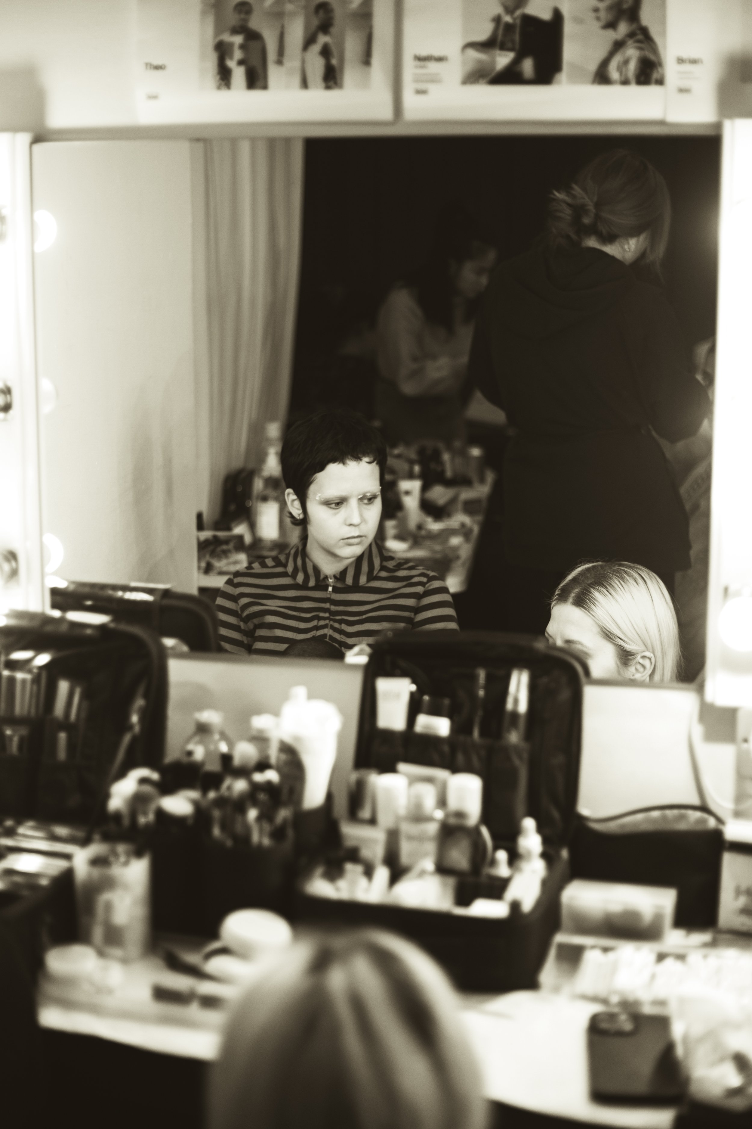 A young person sitting at a makeup station, looking into a mirror with makeup kits and tools on the table. Several other people are in the background, one standing with her back turned, and another person is blurred behind them.