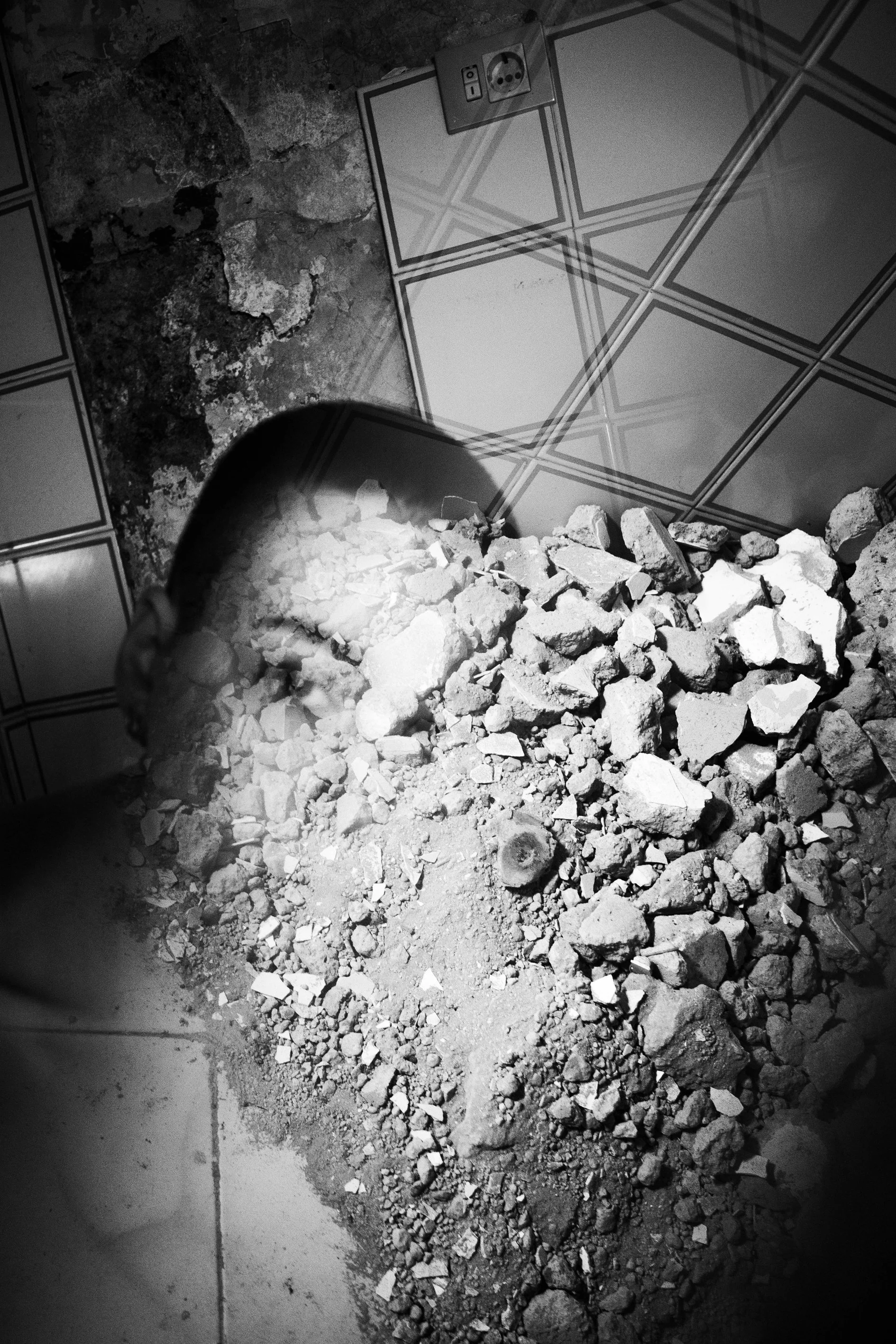 A person with a shaved head sitting on a tiled floor, surrounded by rubble and dirt, with an electrical outlet visible on the wall behind them.