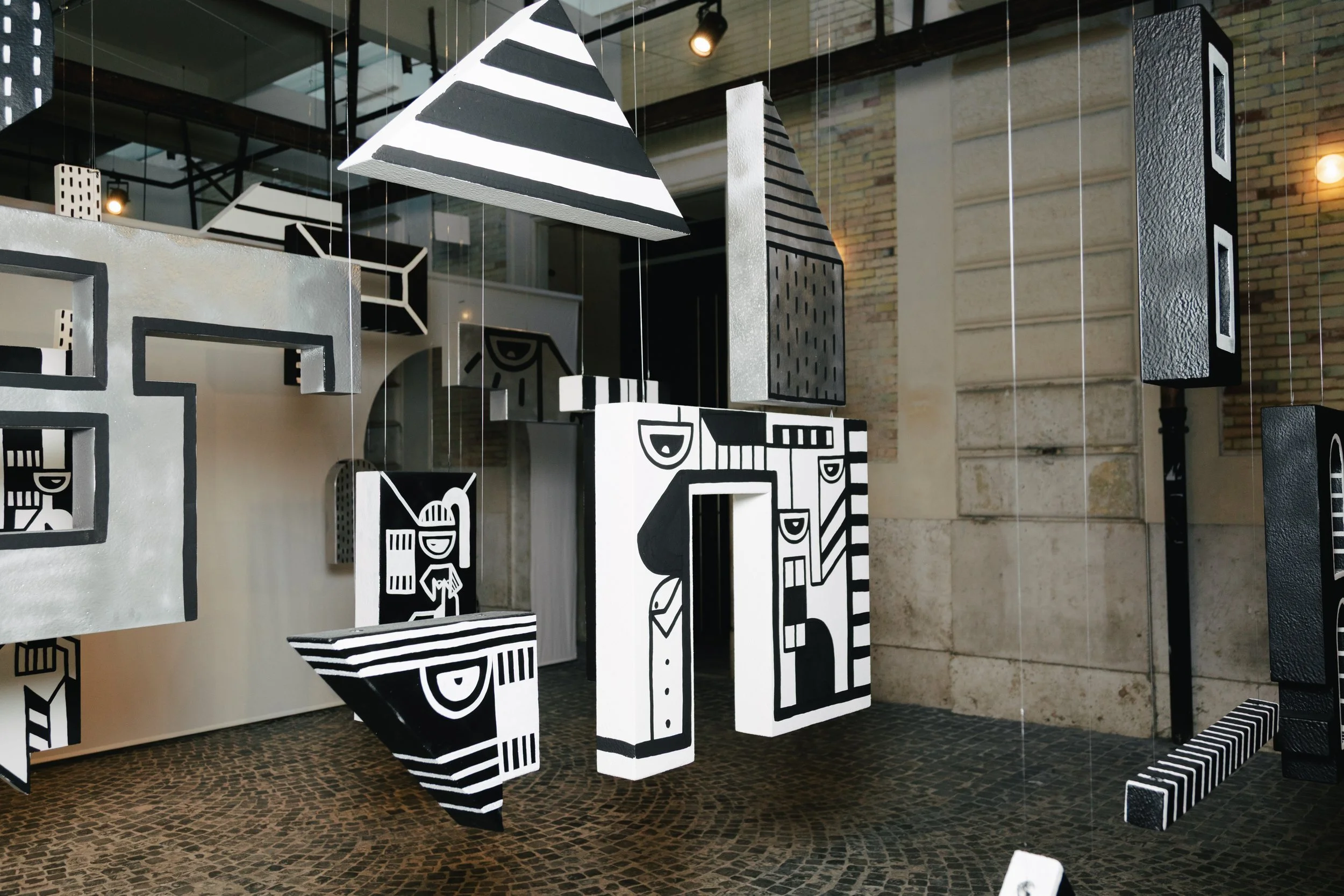 Art installation featuring black, white, and gray geometric and abstract shapes hanging from the ceiling in a gallery space with stone and brick walls.