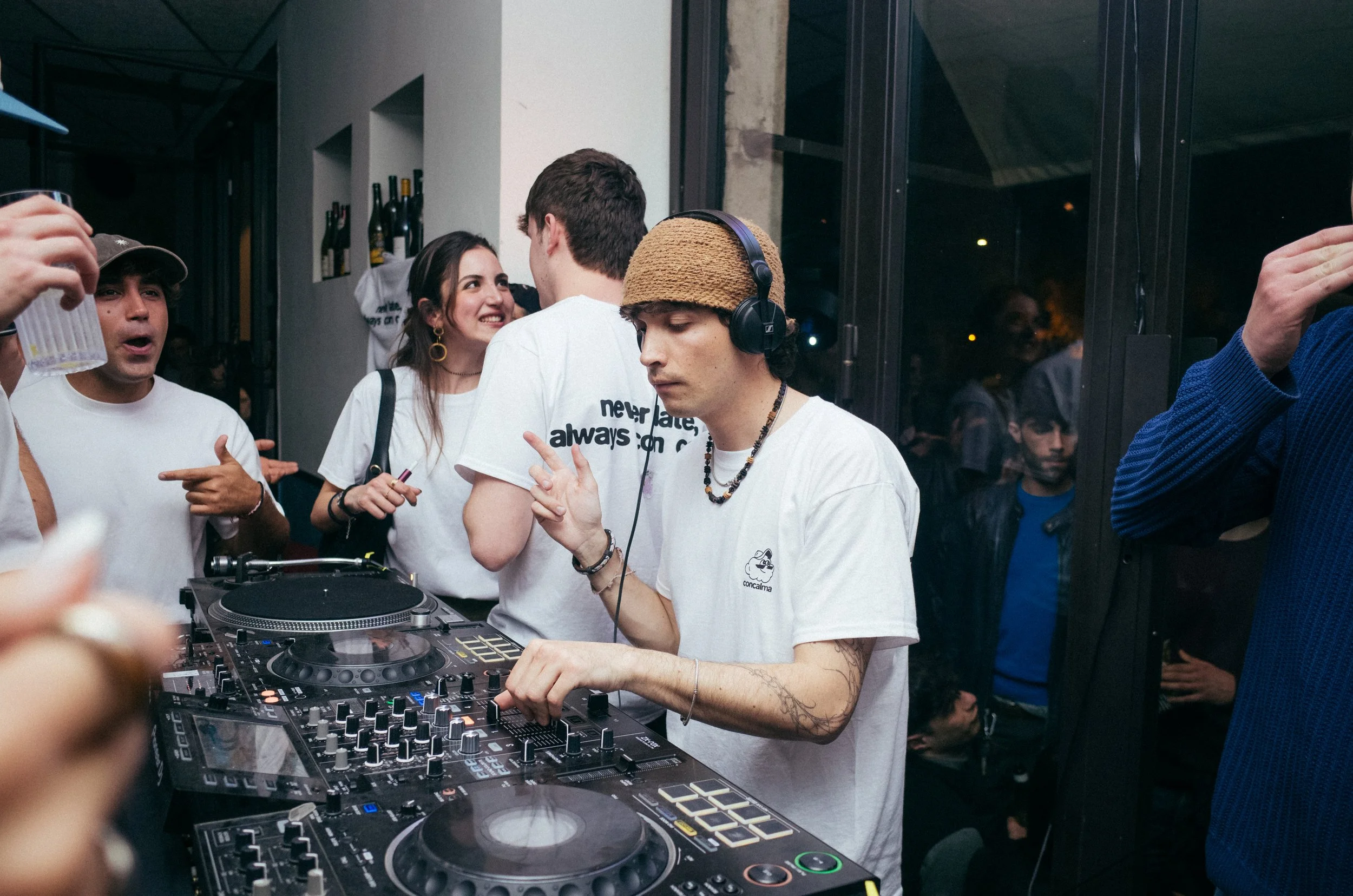 A young male DJ with tattoos, wearing headphones, a knit cap, and a white t-shirt, is operating DJ equipment at a crowded party or club. People around him are engaged, some dancing and talking, enjoying the music.