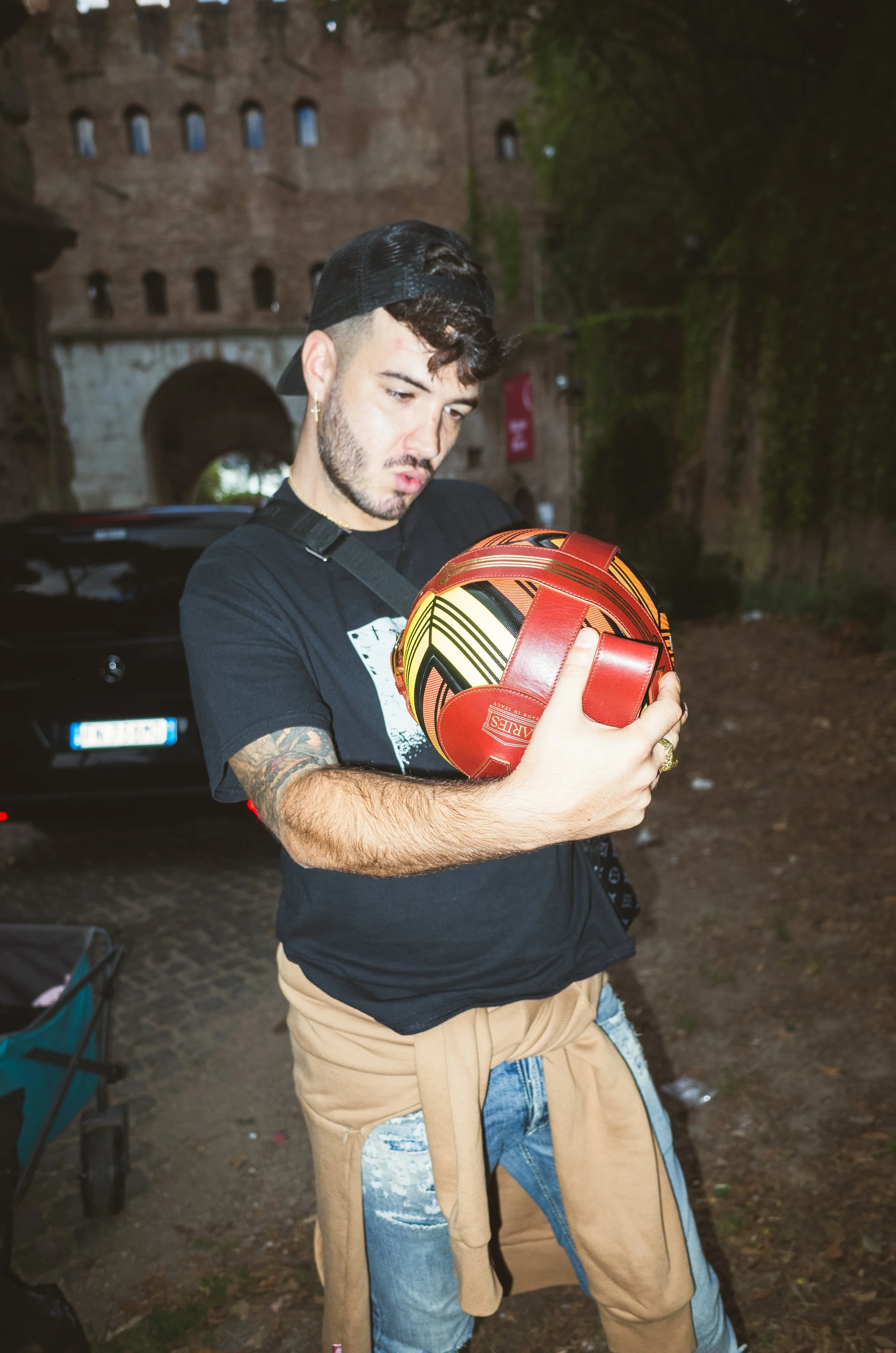 A young man with tattoos, wearing a black t-shirt, tan jeans, and a black cap worn backwards, is holding a red helmet with black and yellow accents, looking at it with a surprised or curious expression. He is outdoors near an old brick structure and 