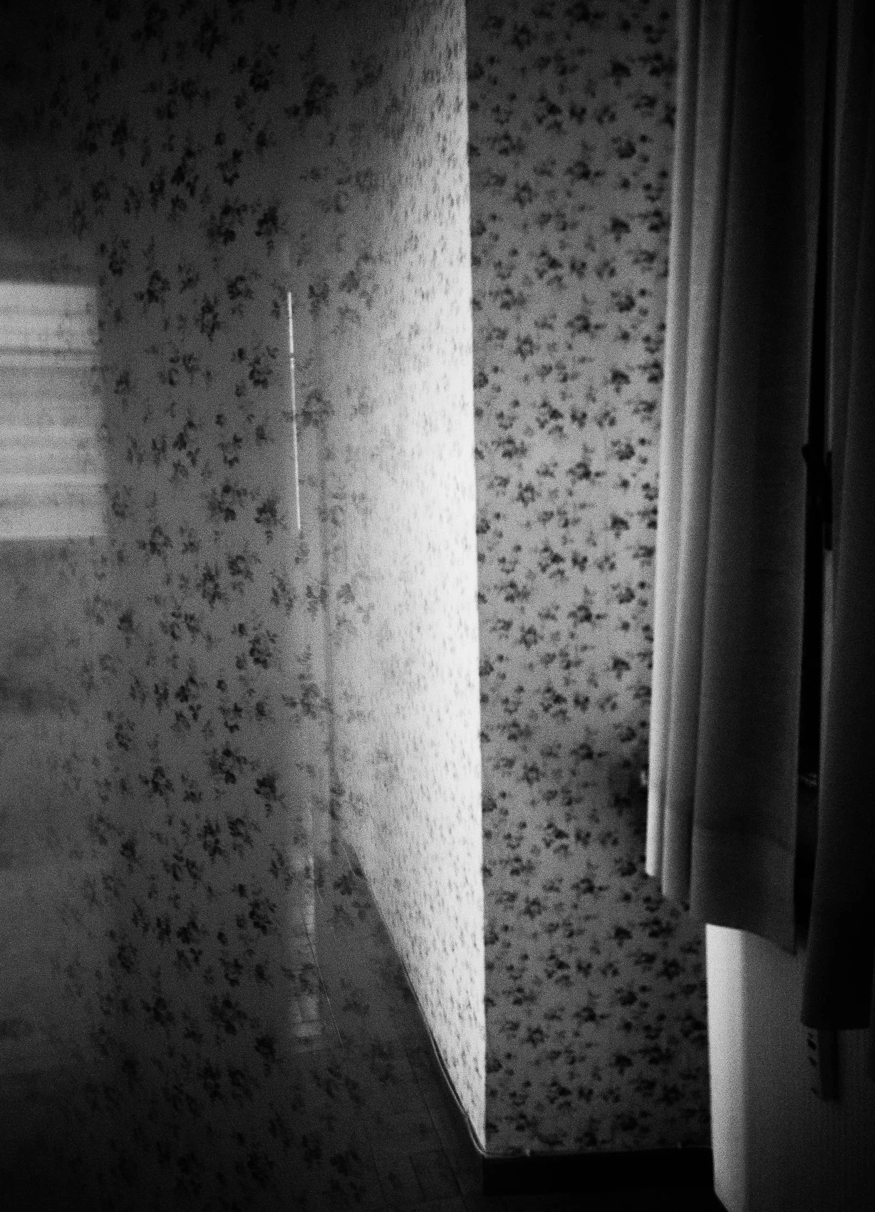 Black and white photo of a corner of a room with floral wallpaper, a door partially open with curtains pulled to the side, and a wooden floor.