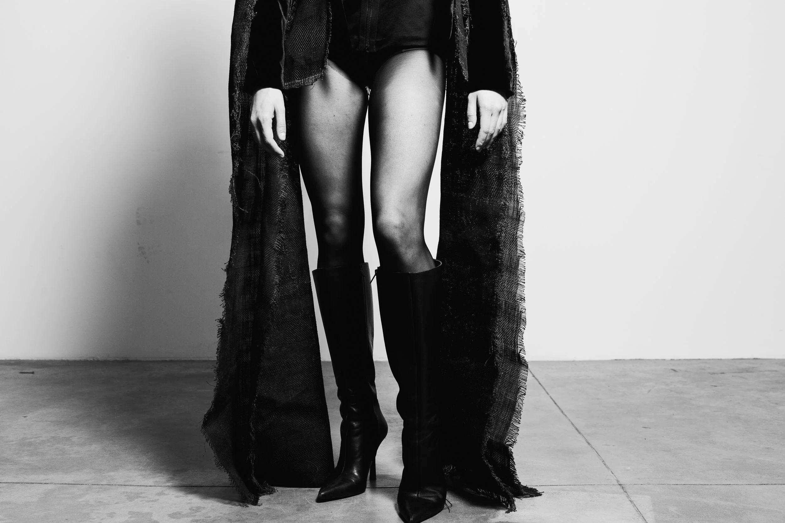 Black and white photo of a woman wearing thigh-high black boots, black shorts, and a long, ripped black coat, standing against a plain wall.