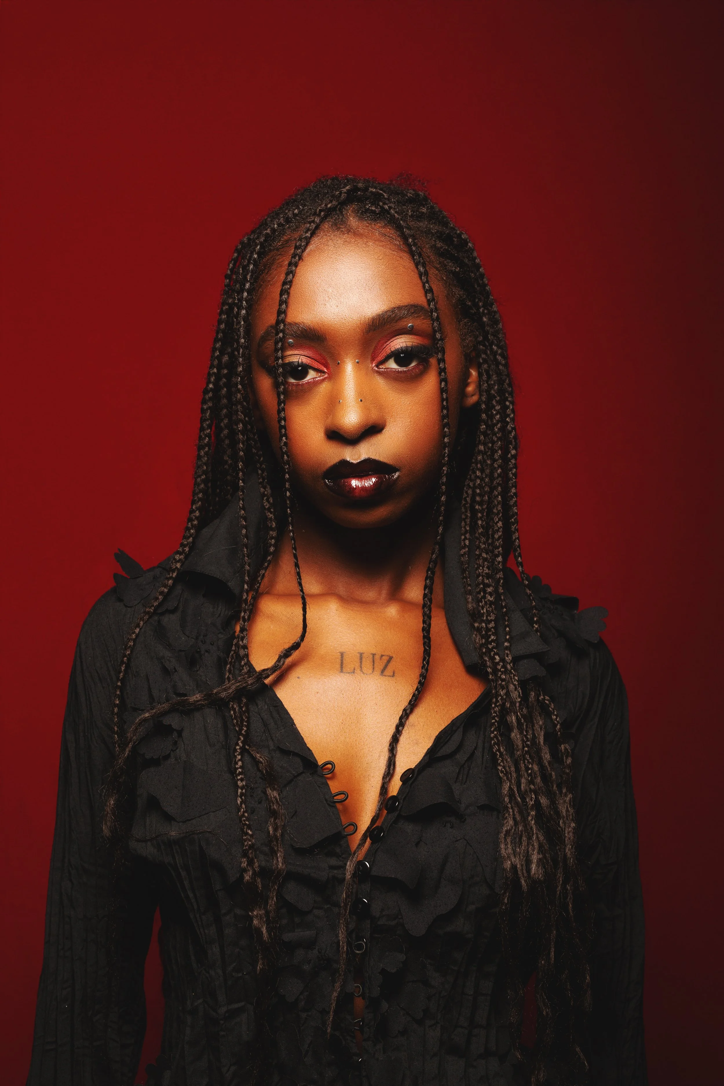 A woman with dark skin and long braids wearing black clothing with ruffles, standing against a red background.